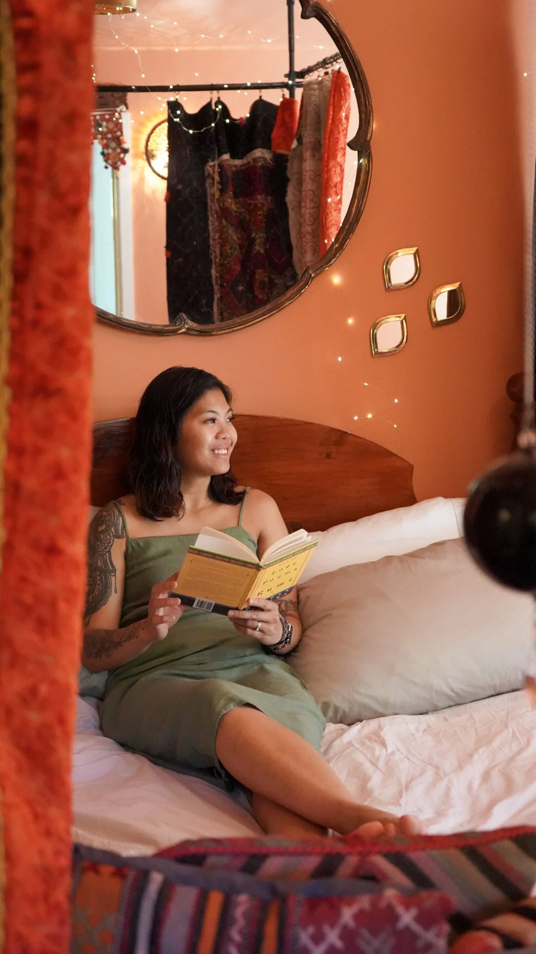 A woman with dark hair and tattoos on her arm is sitting on a bed, holding and reading a yellow book, smiling, in a cozy room with pink walls, a wooden headboard, decorative mirrors, and hanging clothes reflected in a mirror.