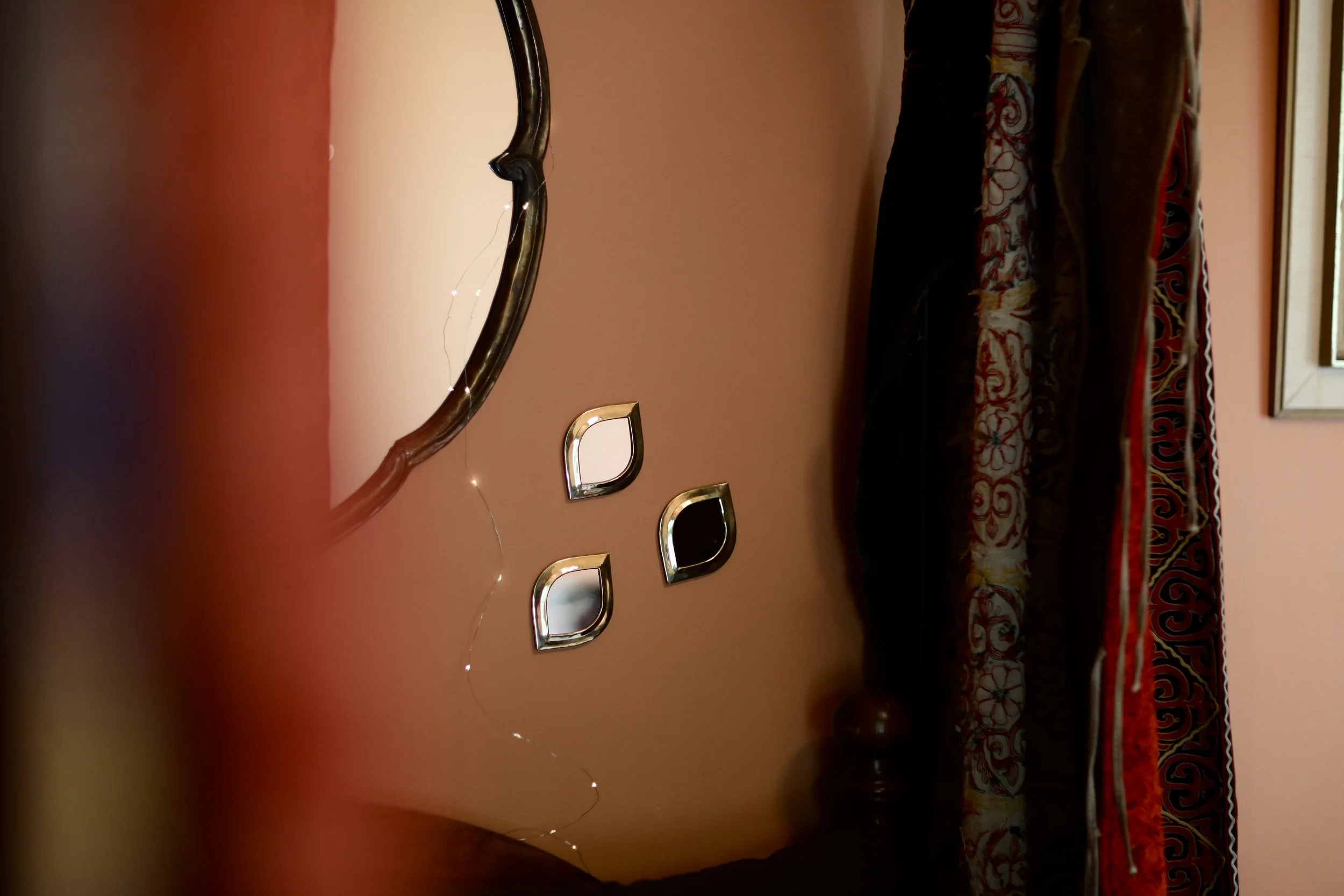 A wall with a mirror, string lights, and three small mirrors, with a bed and colorful bedspread on the right side.