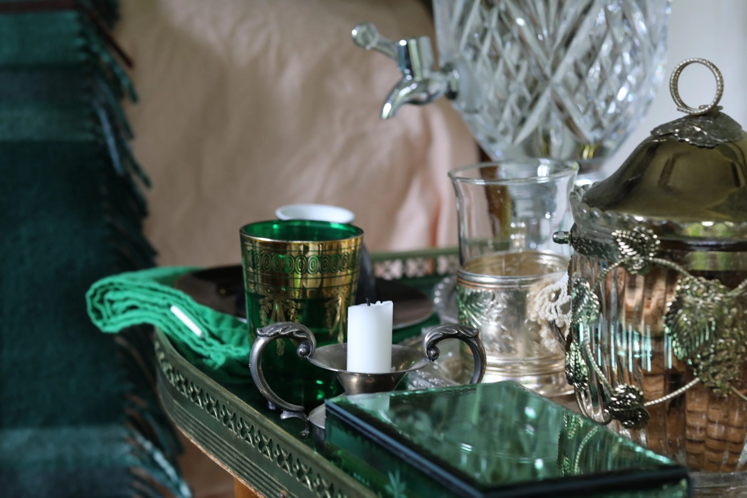 Decorative glassware and table centerpiece on a silver tray, including a white candle, green glass, and silver jewelry, on a table with a green cloth and fringed blanket.