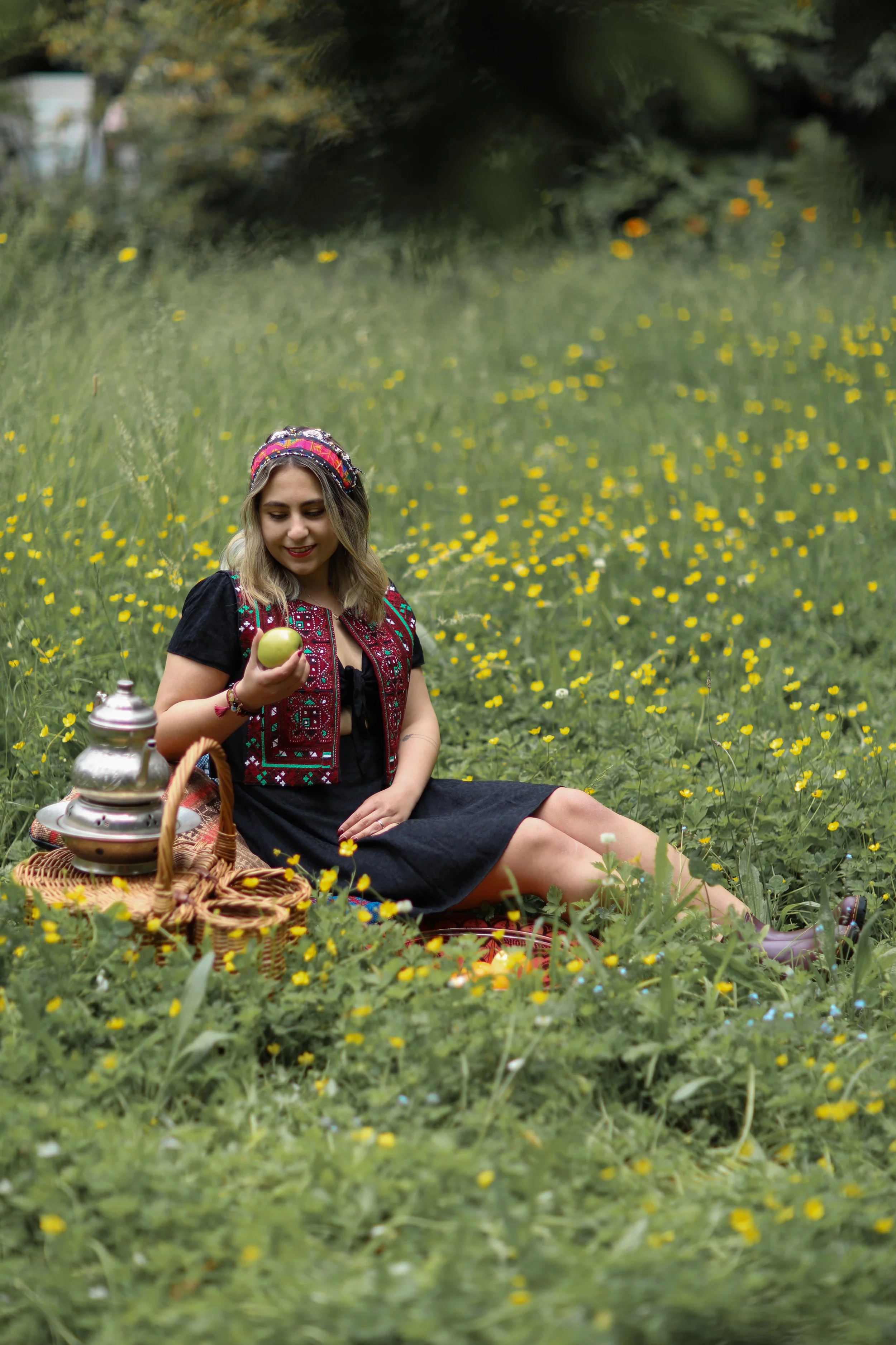 A young woman with blonde hair, wearing traditional folk attire, sitting in a grassy field filled with yellow flowers, holding a green fruit, with a picnic basket and a lantern nearby, under green trees.
