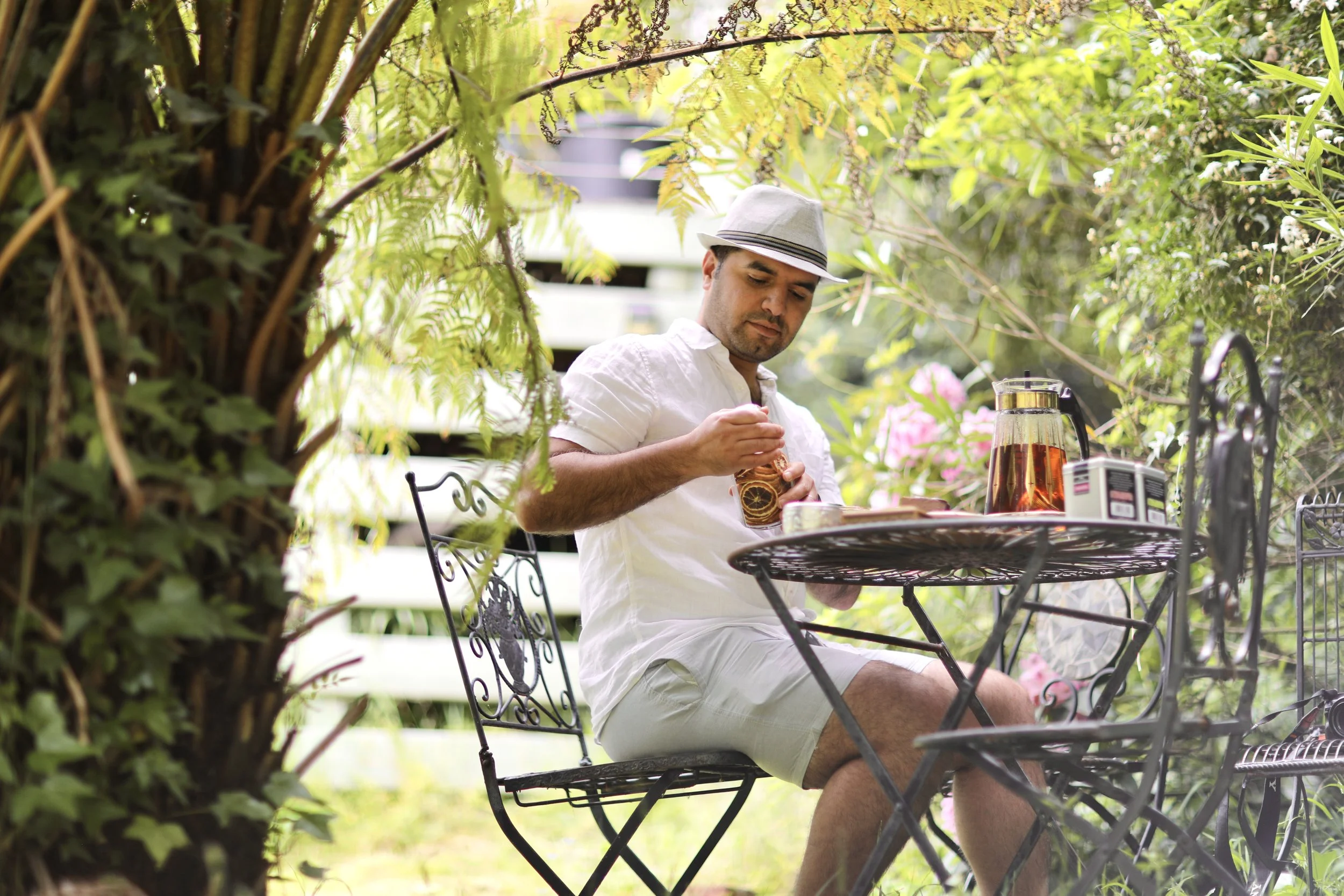 A man sitting outdoors at a small round metal table, wearing a white shirt, white shorts, and a gray fedora hat, holding a drink with a lemon slice, with a pitcher of tea on the table, surrounded by lush green foliage and flowering plants.