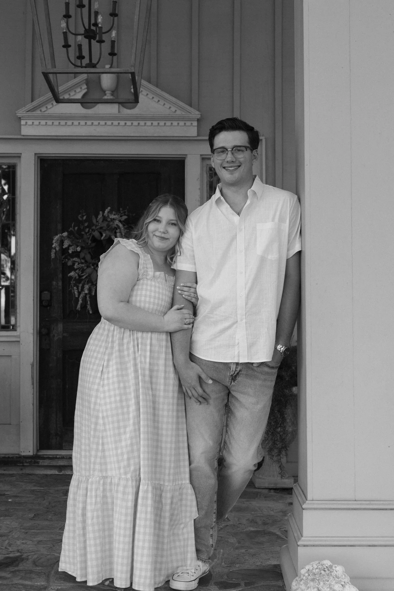 A black and white photograph of a young man and woman standing together in front of a house entrance. The woman is wearing a plaid dress and has her arm linked with the man, who is wearing a short-sleeved shirt and jeans. They are smiling at the came