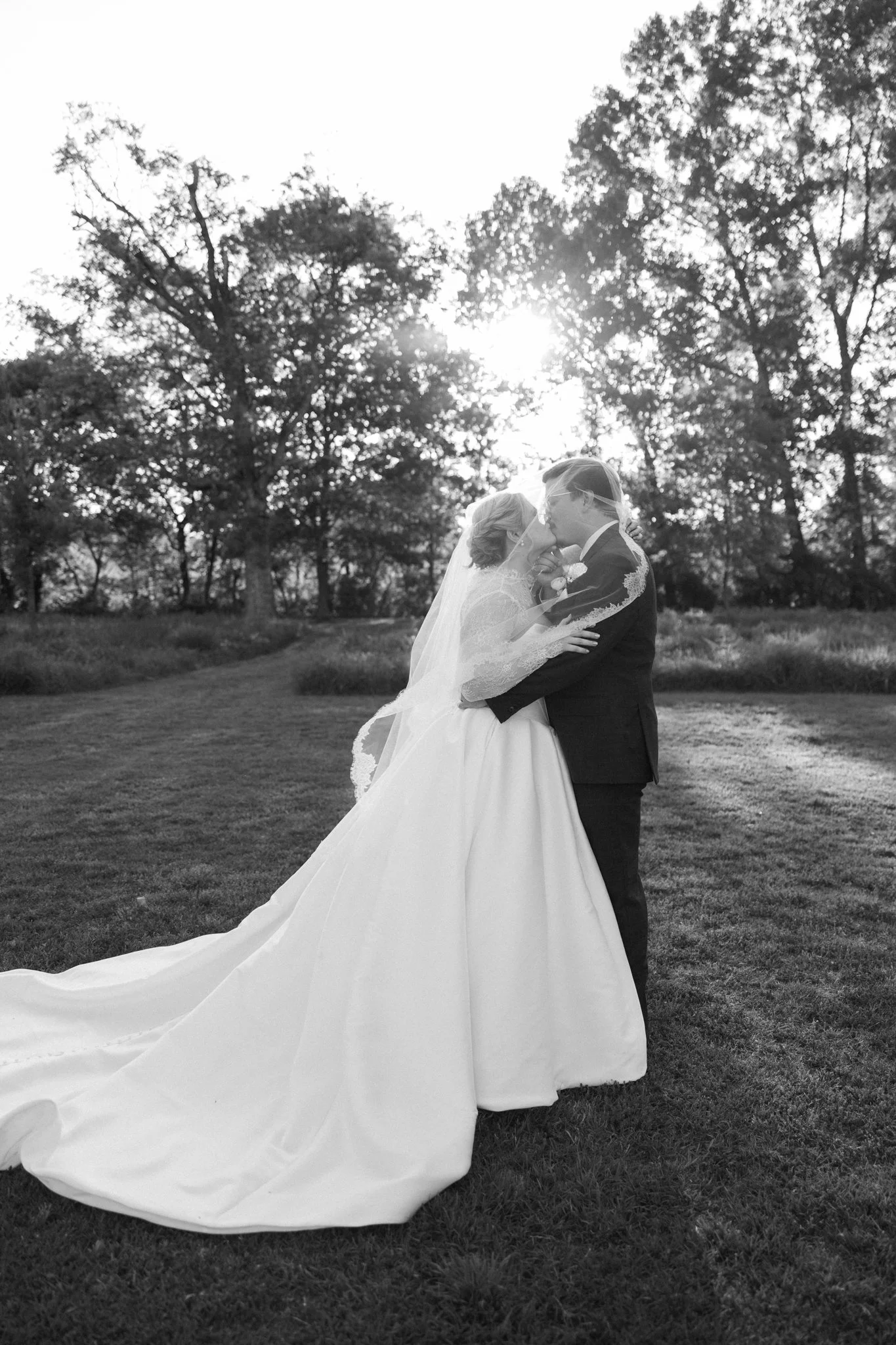 A bride and groom kissing outdoors in a grassy area with tall trees in the background. The bride is wearing a gown with a long train and a veil, and the groom is in a suit. The sun is shining through the trees.