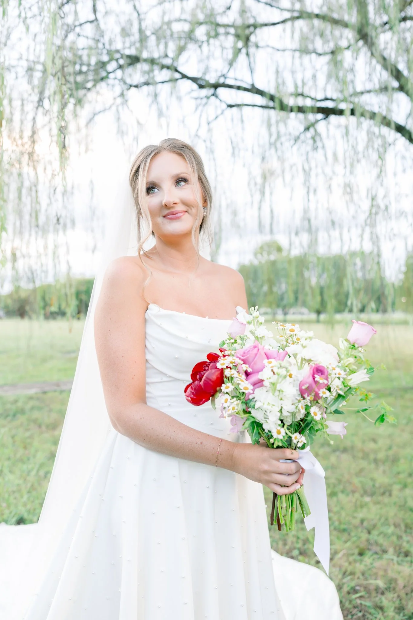 A bride holding a bouquet of pink, red, and white flowers outdoors, standing in front of a weeping willow tree.