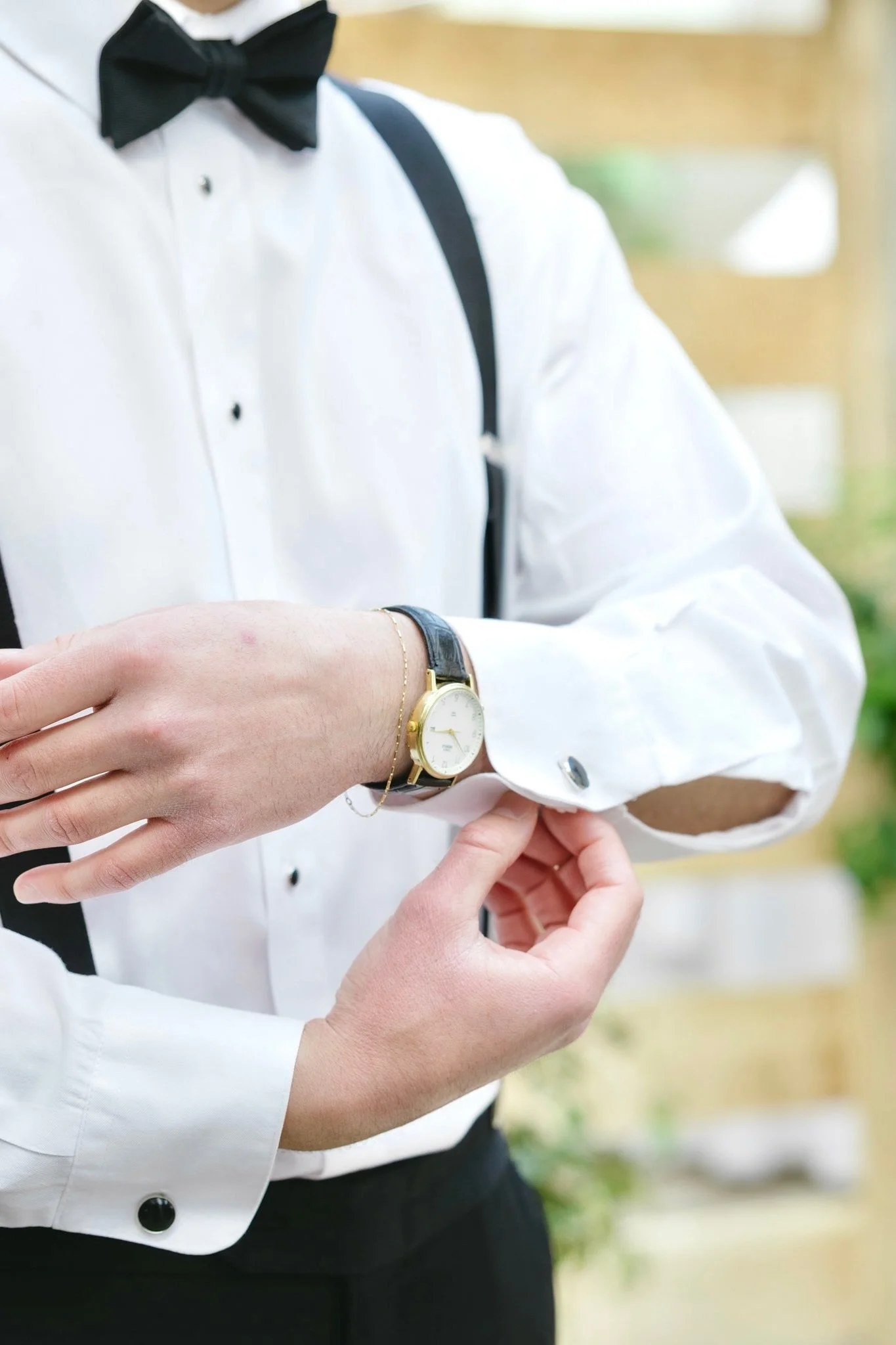 A person in formal attire, wearing a white shirt with a black bow tie, is having their left wrist checked by another individual. The person being checked is wearing a gold wristwatch with a black strap and a delicate bracelet, and has a black shoulder bag. The background is blurred with hints of greenery and natural outdoor setting.