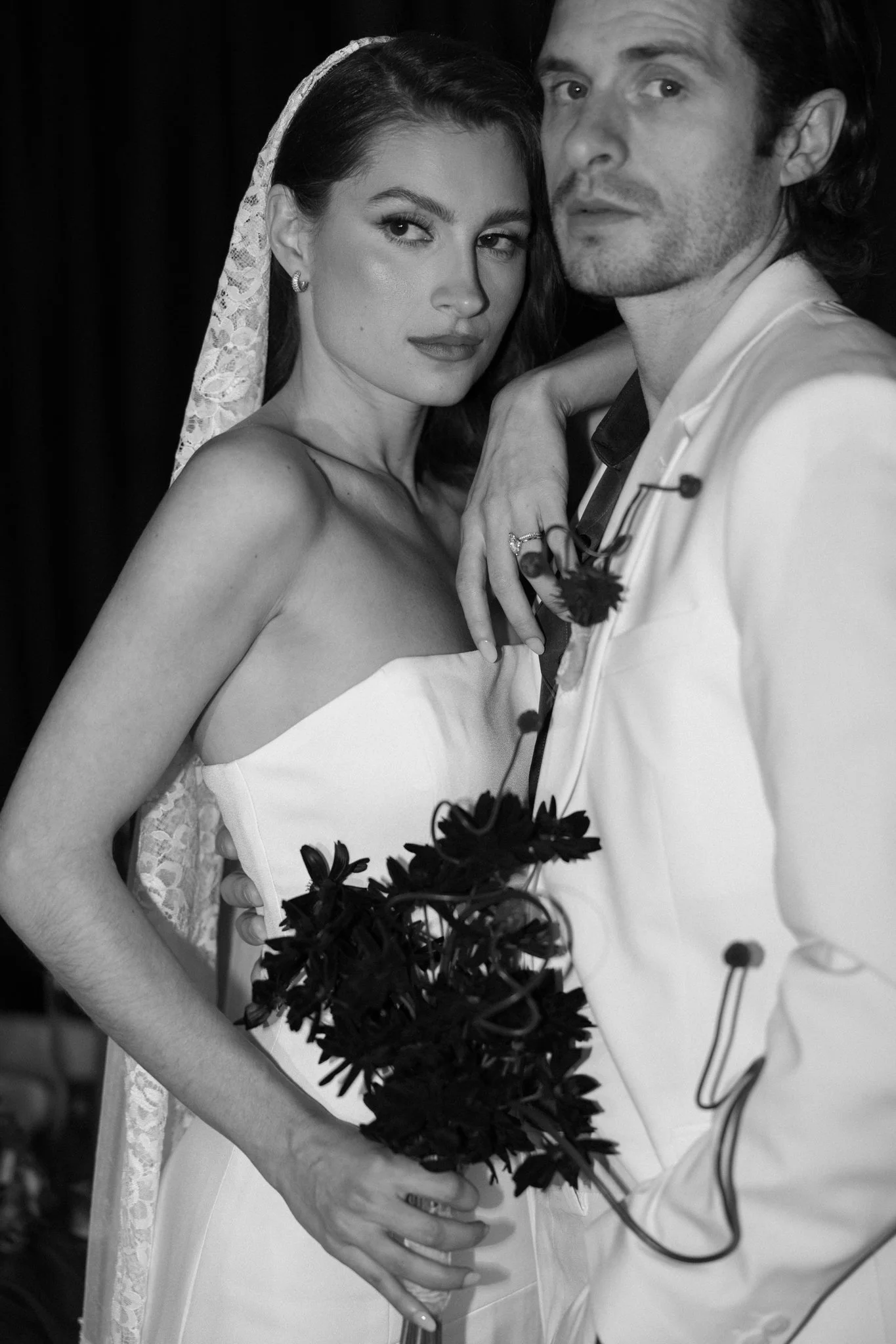 A black and white photo of a bride and groom close together, with the bride wearing a wedding dress and lace veil, and the groom in a white suit, holding a bouquet of dark flowers.