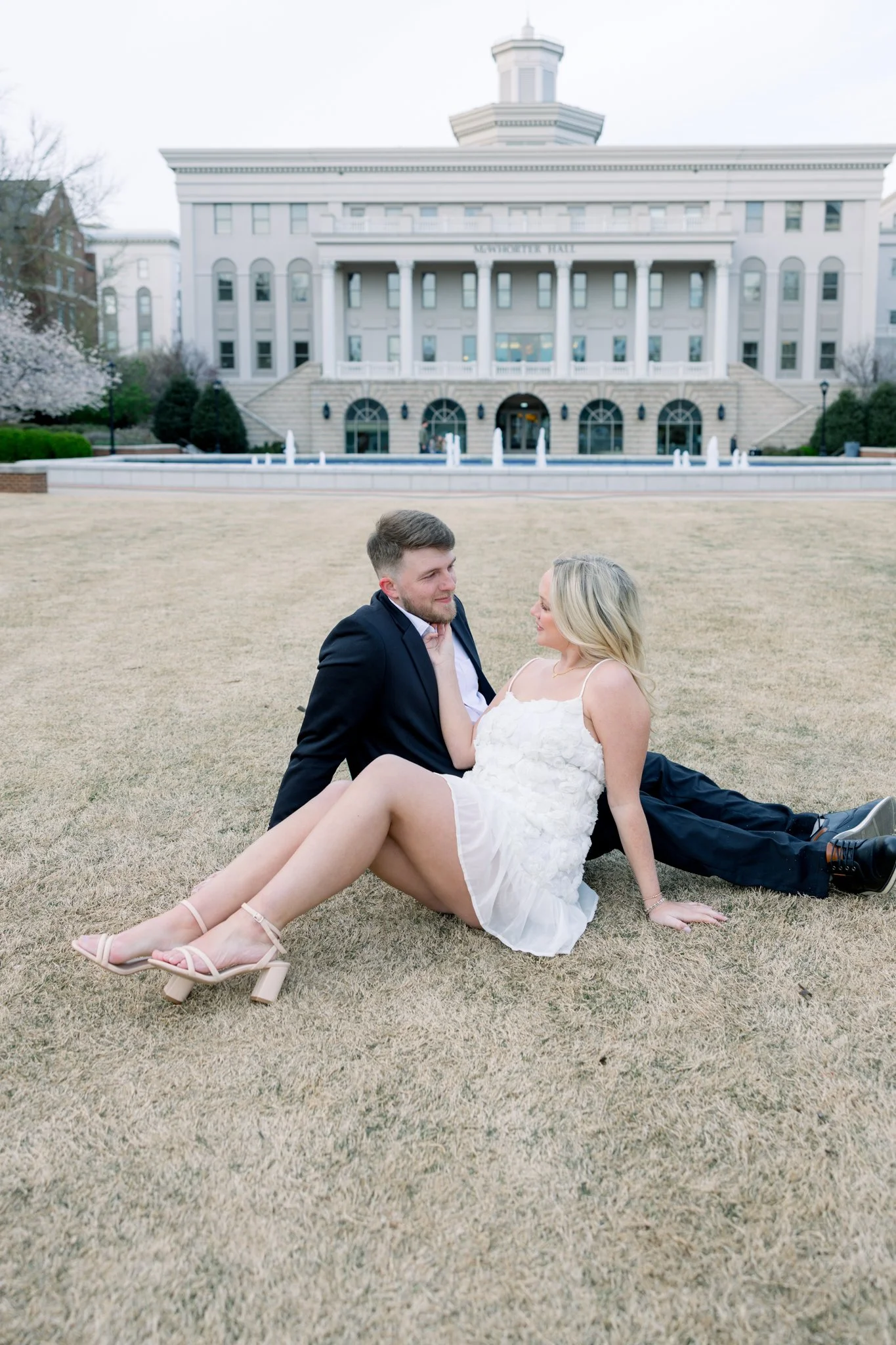 A man and woman sitting closely on a patchy lawn in front of a large white government building, possibly a courthouse or city hall, with steps and columns, during daytime.