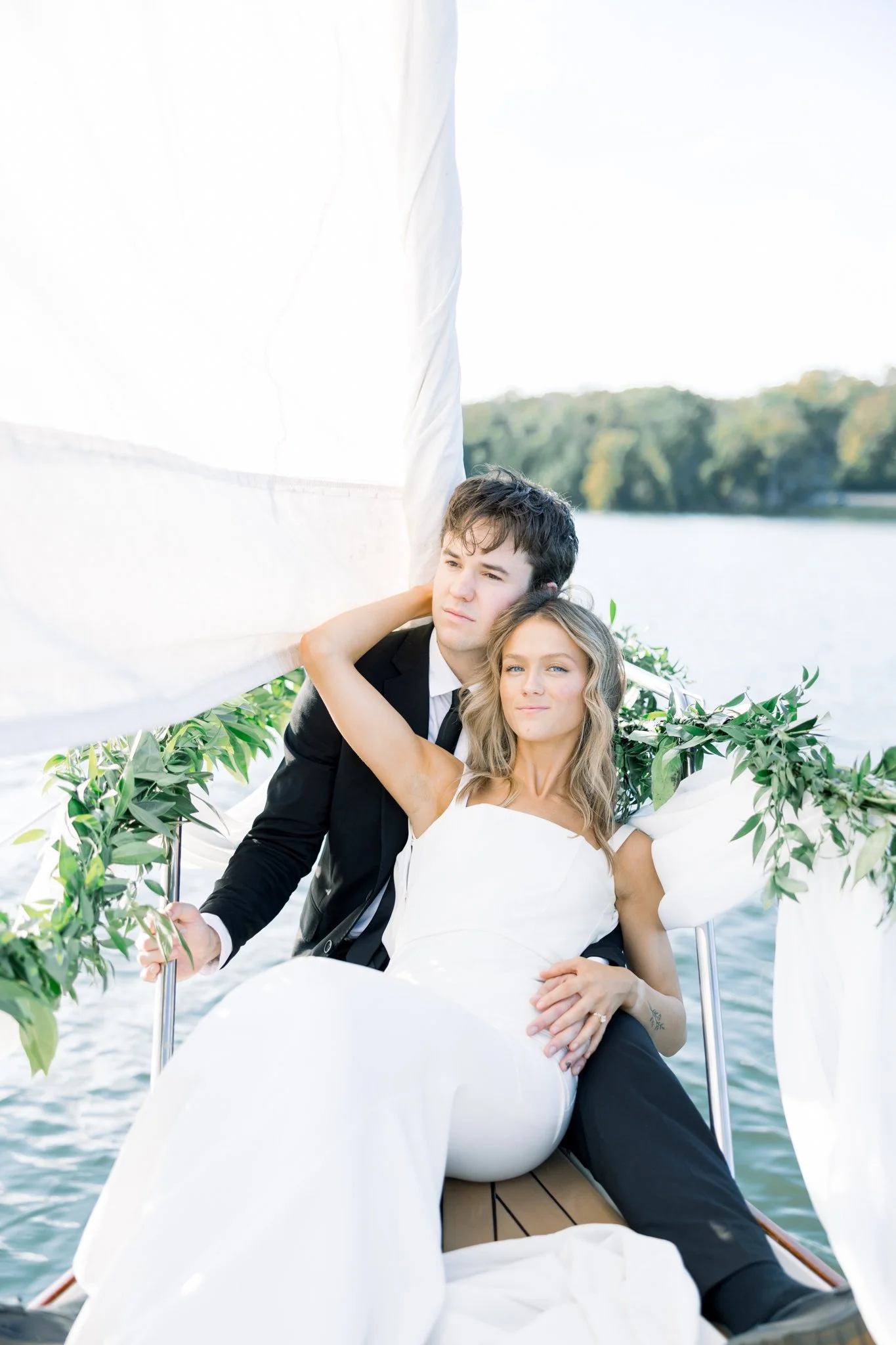 A couple dressed in wedding attire sitting on a boat with a lake and trees in the background.