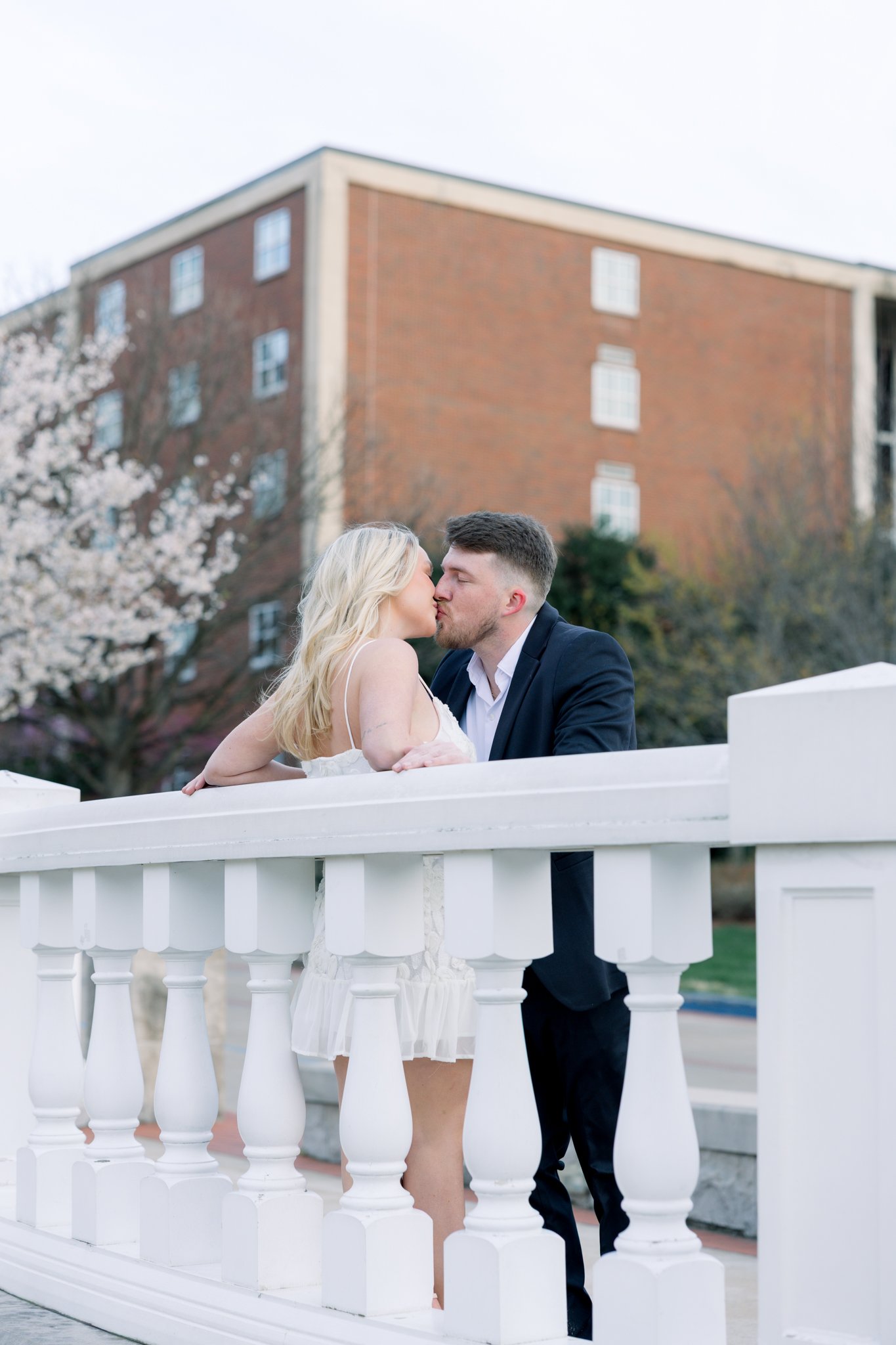 A man and woman kissing on a white bridge outdoors, with a brick building and blooming trees in the background.