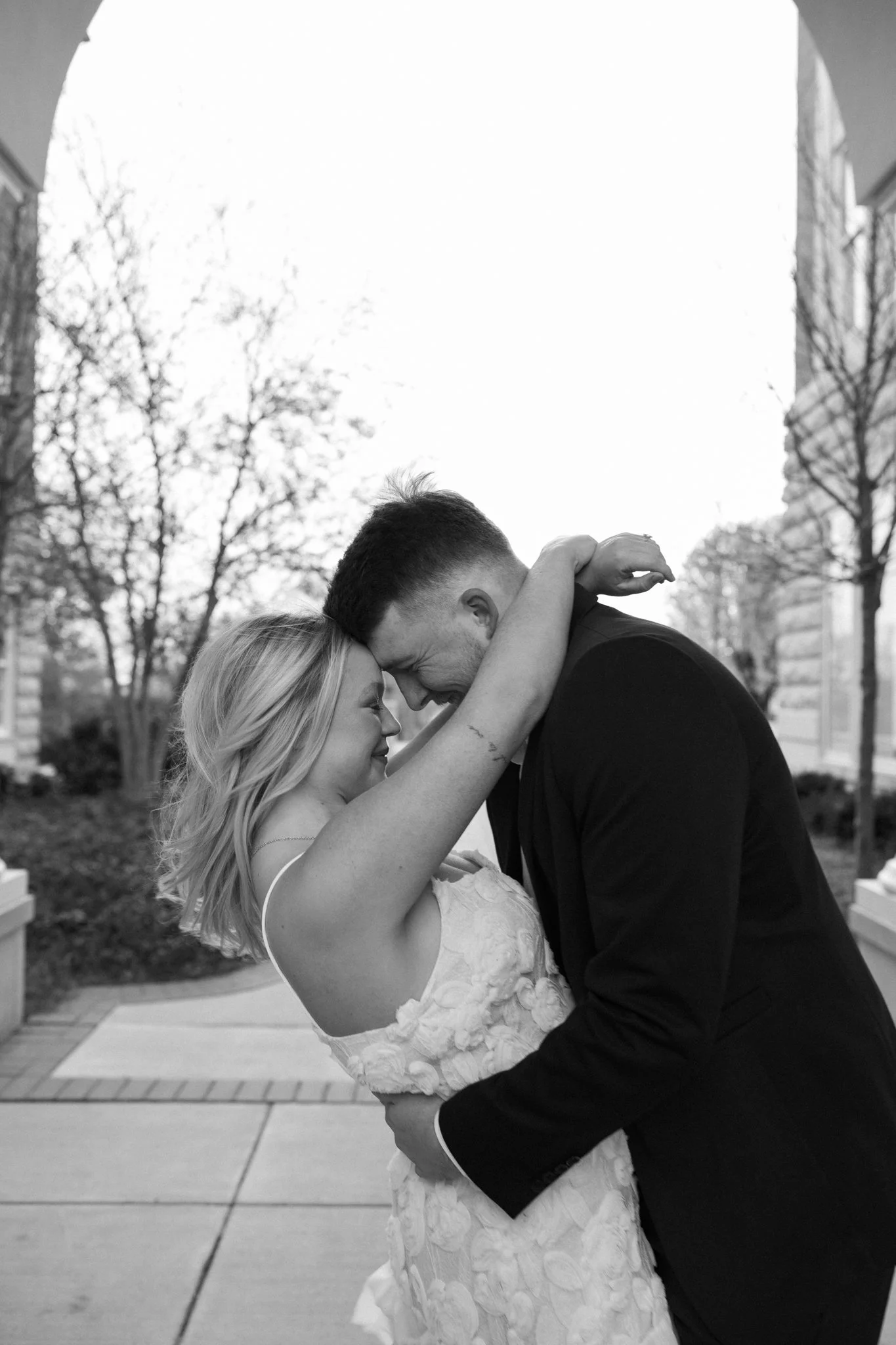 A wedding couple sharing a close, intimate moment outdoors, with the bride dressed in a lace gown and the groom in a black suit, both smiling, surrounded by trees and buildings, in black and white.