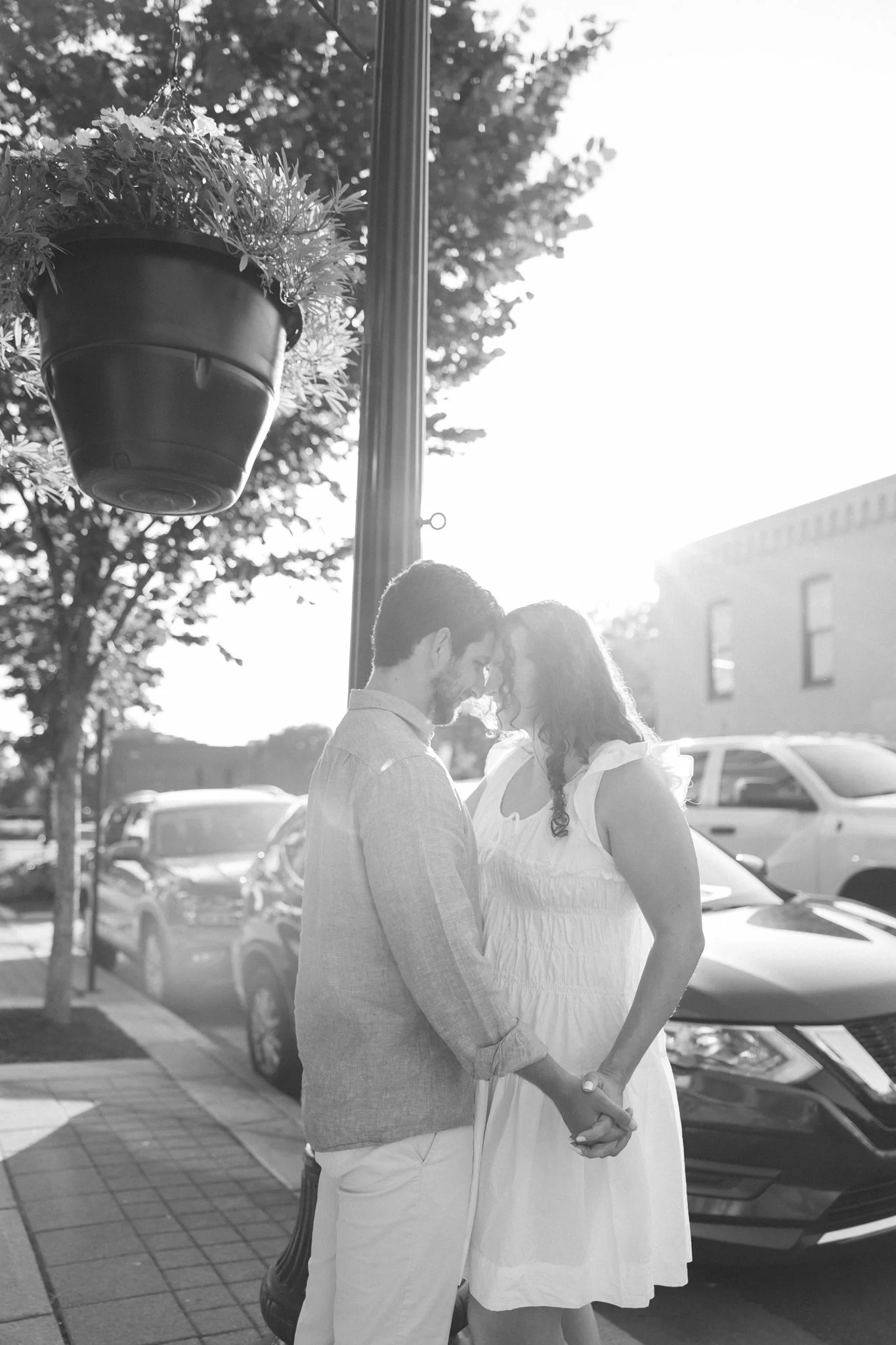 A black and white photo of a couple sharing an intimate moment outdoors, holding hands and touching foreheads, with cars parked in the background.