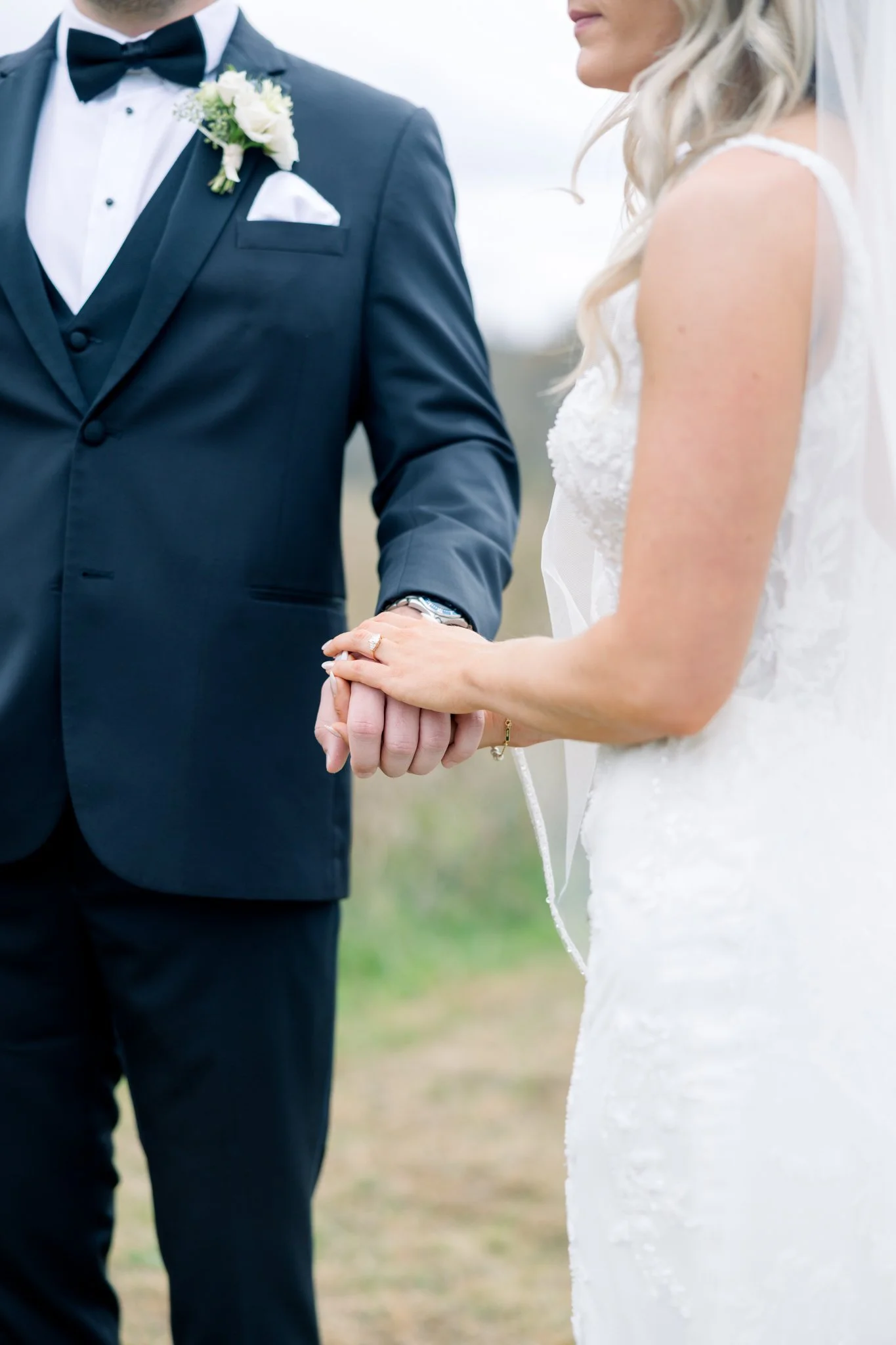 A bride and groom holding hands during a wedding ceremony outdoors, with the groom in a black tuxedo and bow tie, and the bride in a white wedding dress.