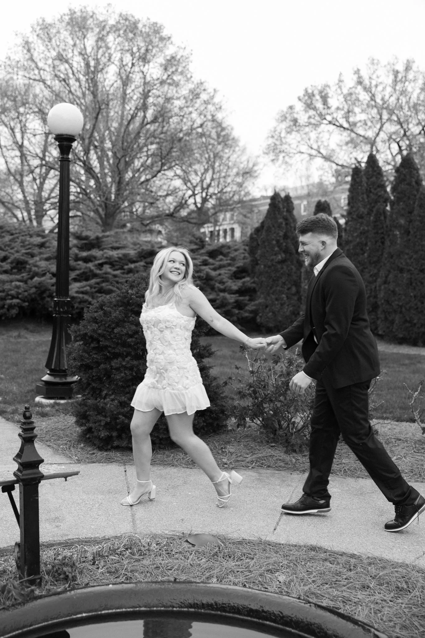 A woman in a dress and high heels dancing outdoors with a man in a suit, smiling and holding hands, surrounded by trees and bushes, with a streetlamp nearby.