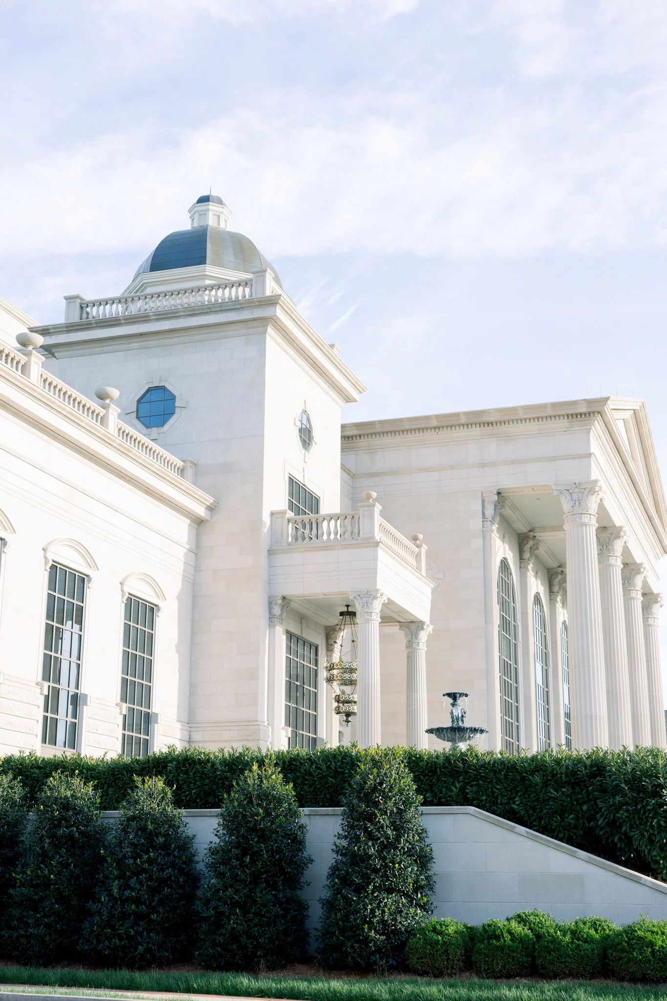 White neoclassical building with columns, a fountain, and greenery in foreground, under a partly cloudy sky.