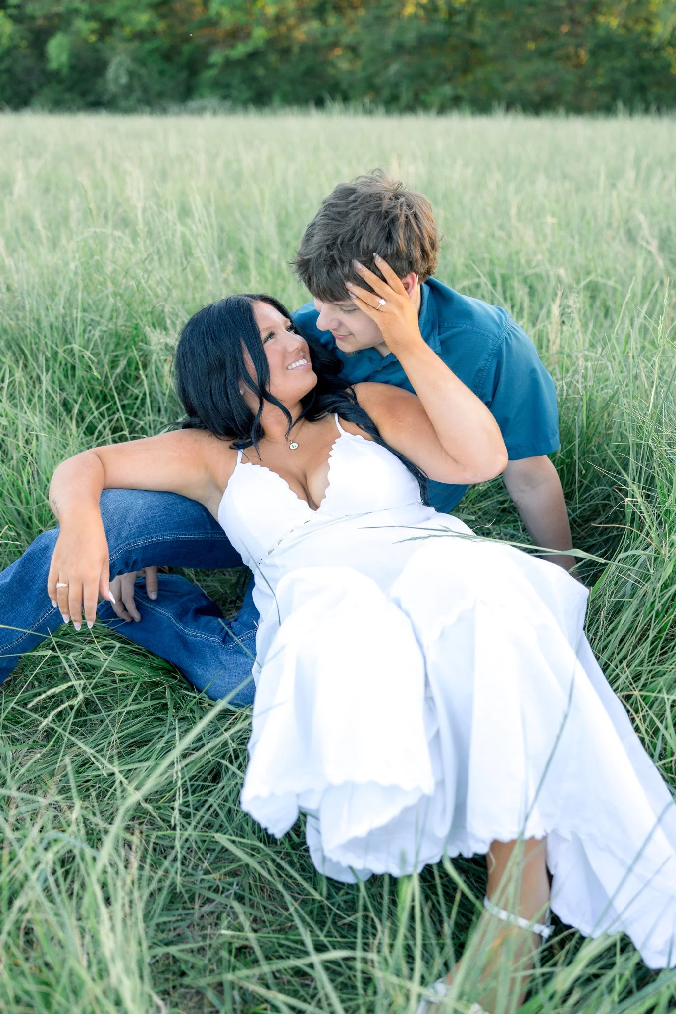 A couple sitting in a grassy field with trees in the background, smiling and looking at each other, with the woman touching the man's face.