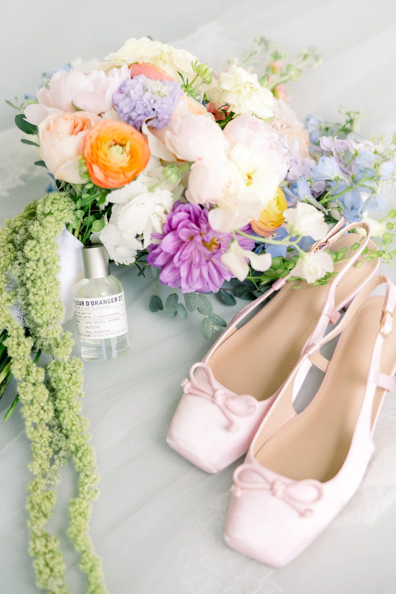 Soft pink ballet flats with ribbons, a bouquet of colorful flowers, and a small perfume bottle on a light surface.