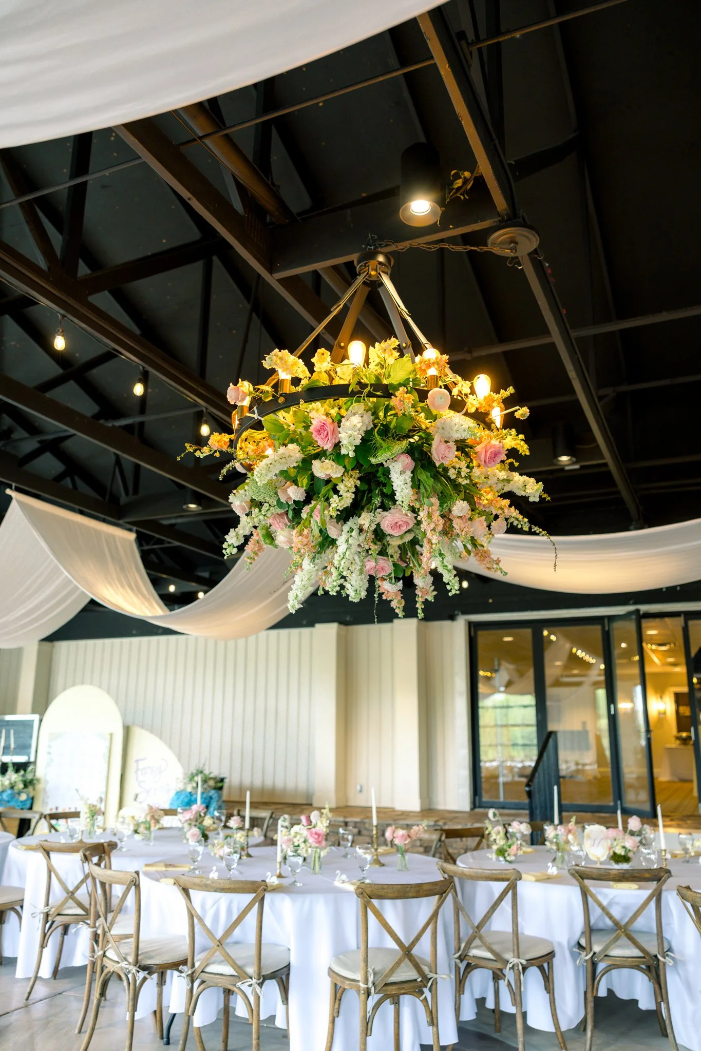 Indoor event space decorated for a celebration with a floral chandelier hanging from the ceiling, tables set with white tablecloths and small flower arrangements, and chairs arranged around the tables.