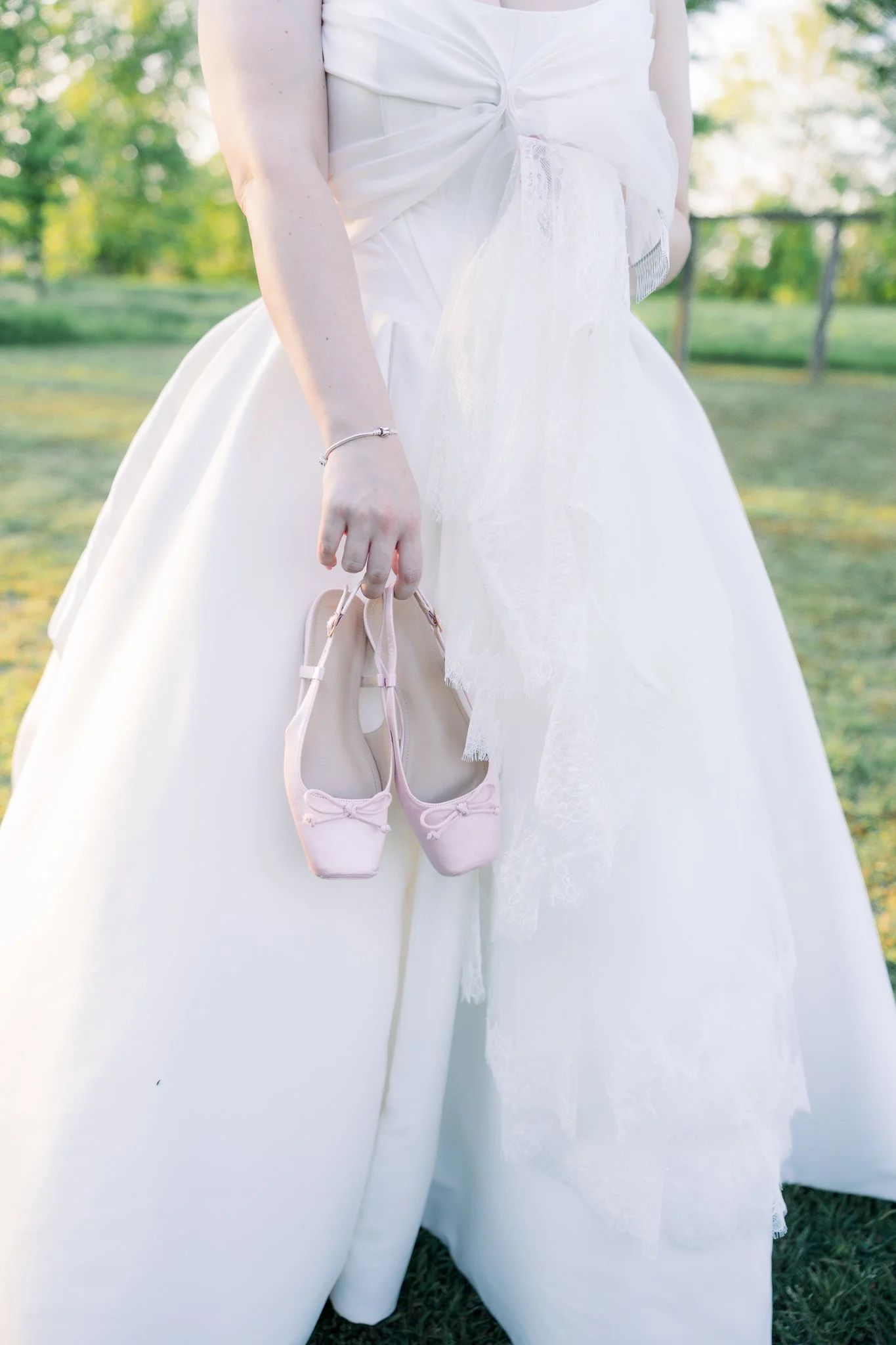 A person wearing a white dress holding pink ballet shoes outdoors with greenery and trees in the background.