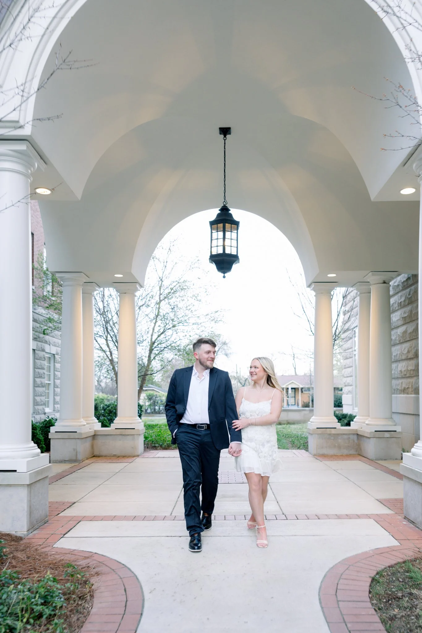 A young couple dressed in wedding attire walking hand in hand beneath a white porch with columns and a hanging lantern, on a cloudy day with leafless trees in the background.