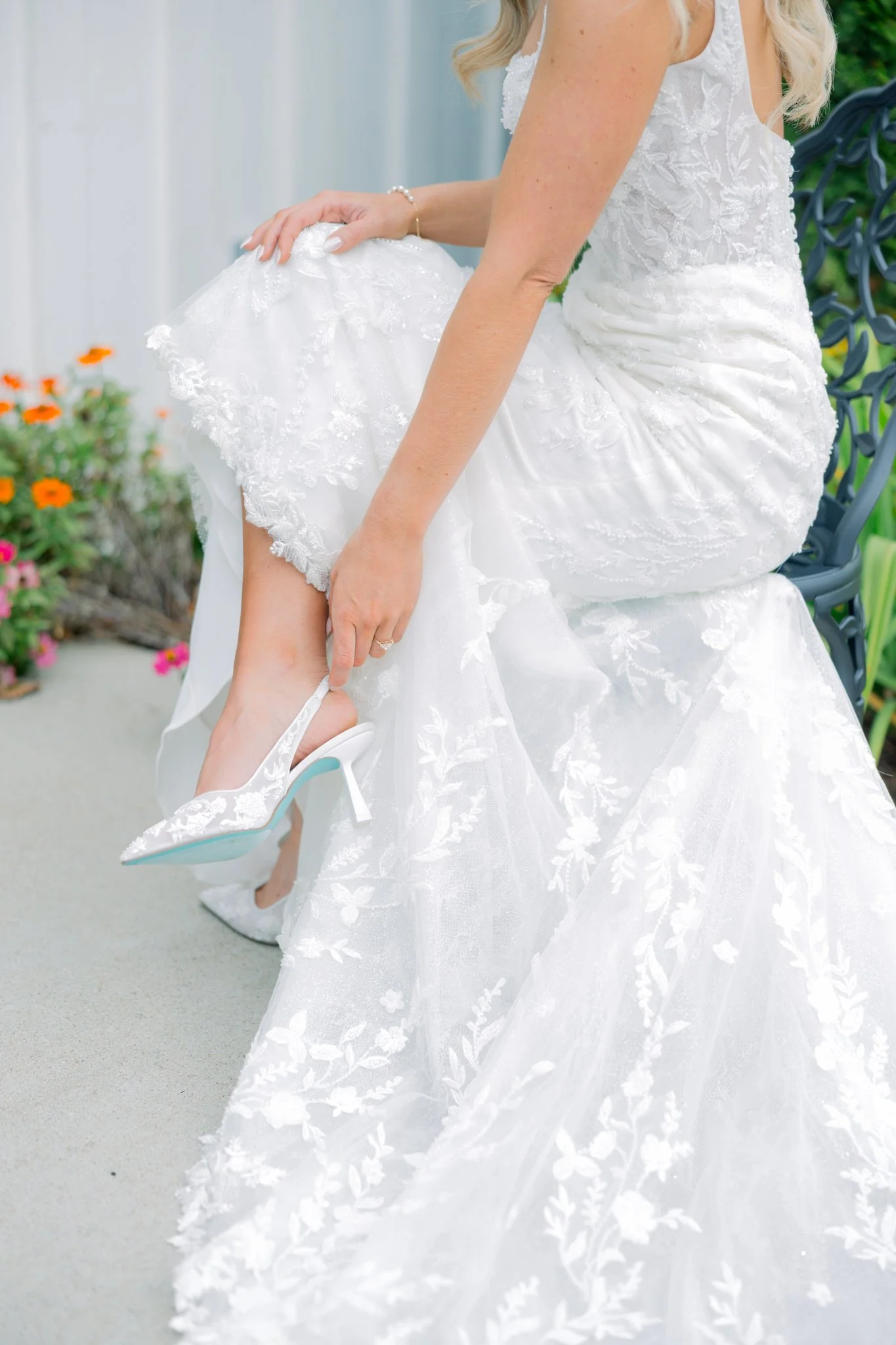 Bride in a white wedding dress adjusting her white high-heeled shoe while sitting on a decorative gray bench outside among orange and pink flowers.