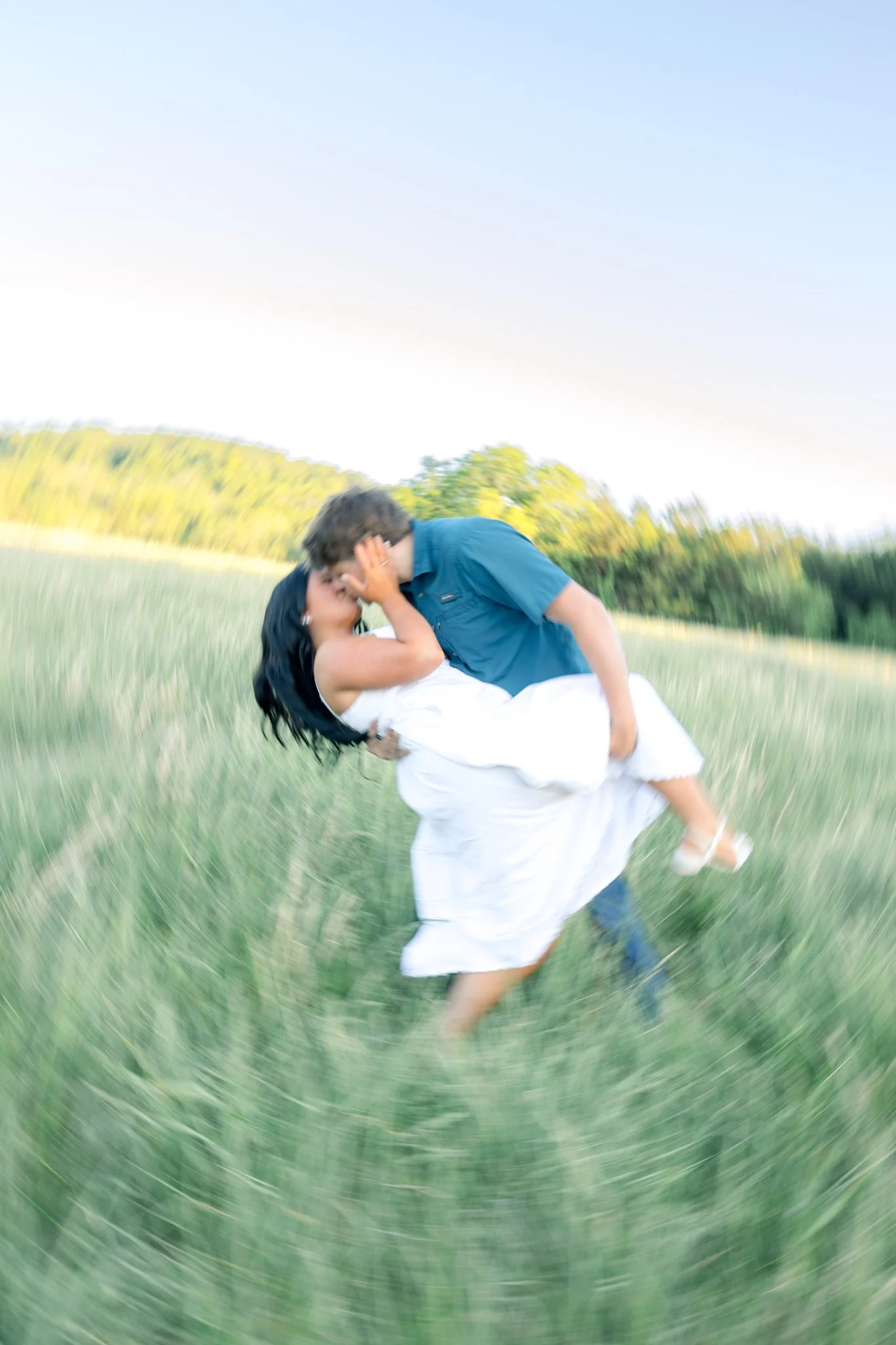 A man holding a woman in a white dress as they kiss in a grassy field with trees in the background during daylight.