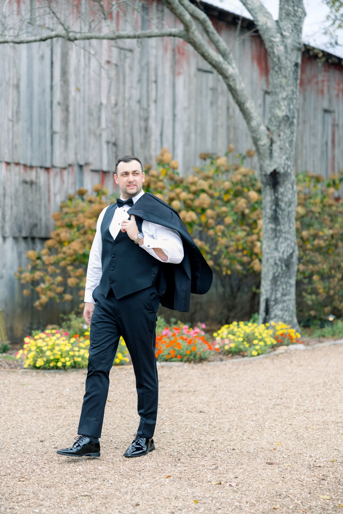 A man in a tuxedo with a bow tie is holding a suit jacket over his shoulder and standing outdoors on a gravel path. Behind him are flowers, a tree, and a weathered wooden fence.
