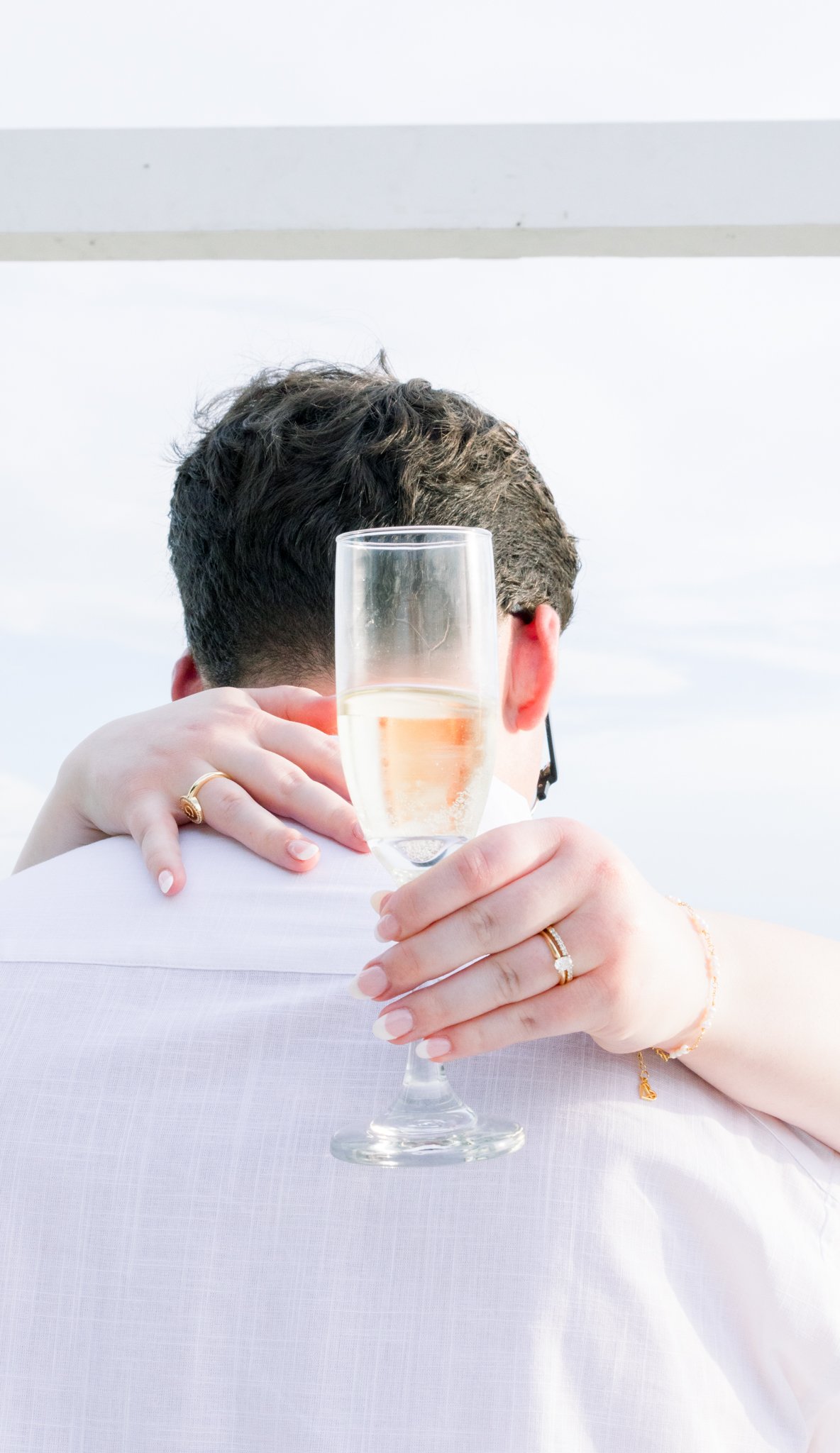 Couple embracing, person holding a glass of champagne in front of the other, focus on wine glass and hands, wedding rings visible, love and celebration mood, outdoor setting with bright sky.