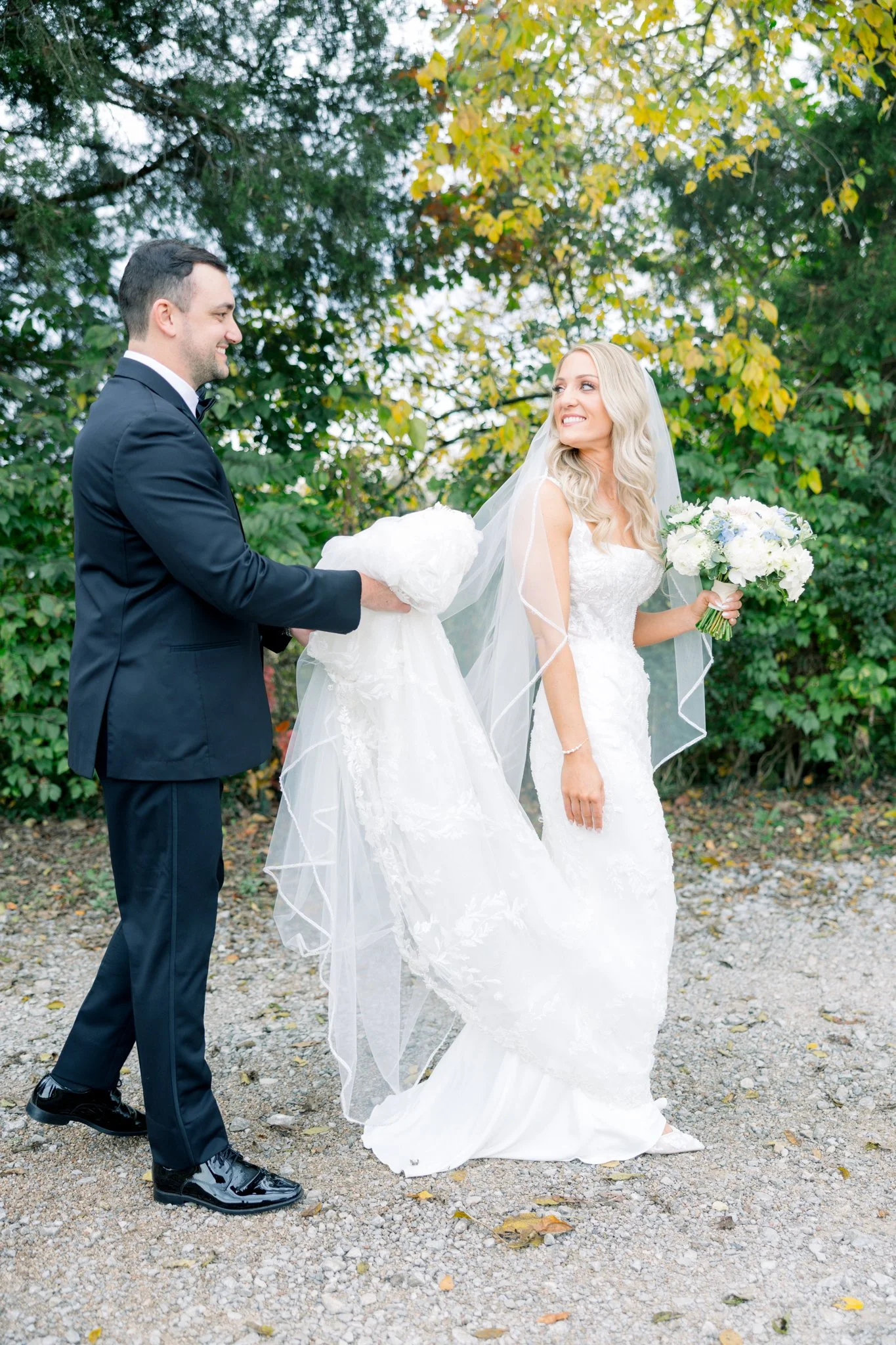A bride and groom smiling at each other outdoors during a wedding photo shoot, with green and yellow foliage in the background.
