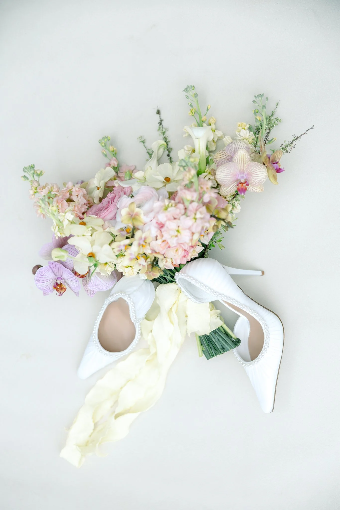 White high-heeled shoes with pearl accents, a cream-colored ribbon, and a bouquet of pink, white, and purple flowers with green foliage, all set against a plain white background.