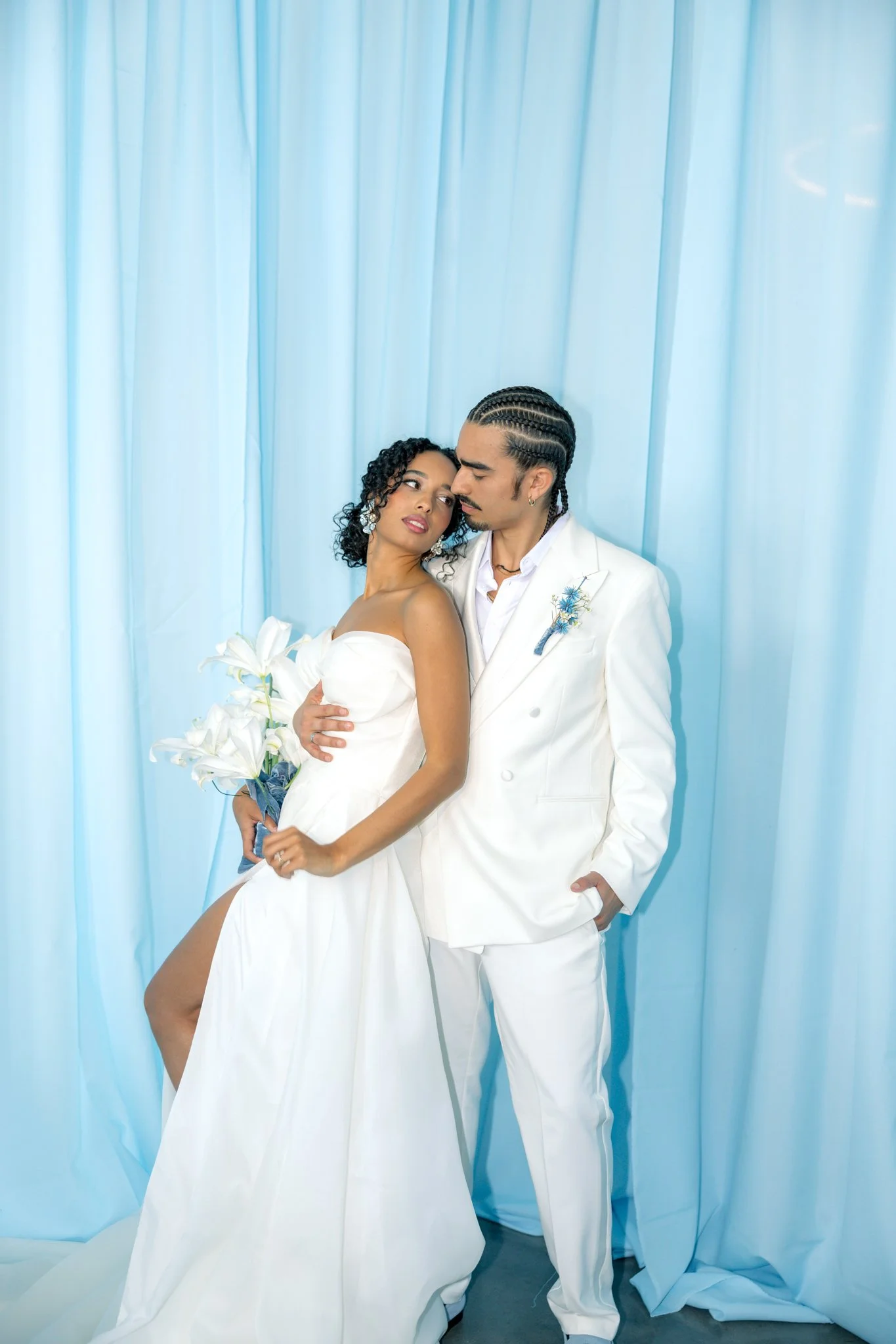 A couple dressed in white wedding attire posing in front of a light blue curtain backdrop. The woman has curly hair and is holding a bouquet of white lilies, while the man has braided hair and is wearing a white suit with a flower boutonniere.