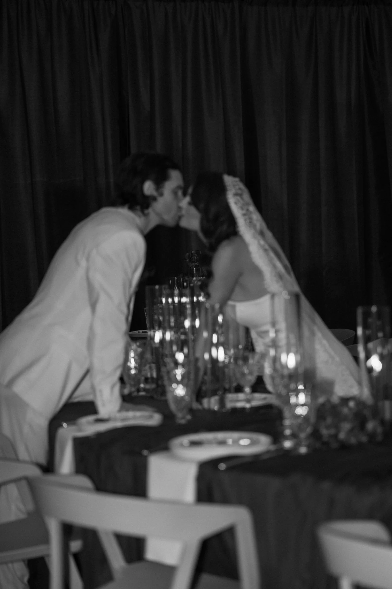 A black and white photo of a man and woman sharing a kiss at a dining table in a restaurant or event setting with dark curtains in the background. The table has multiple glasses and plates.
