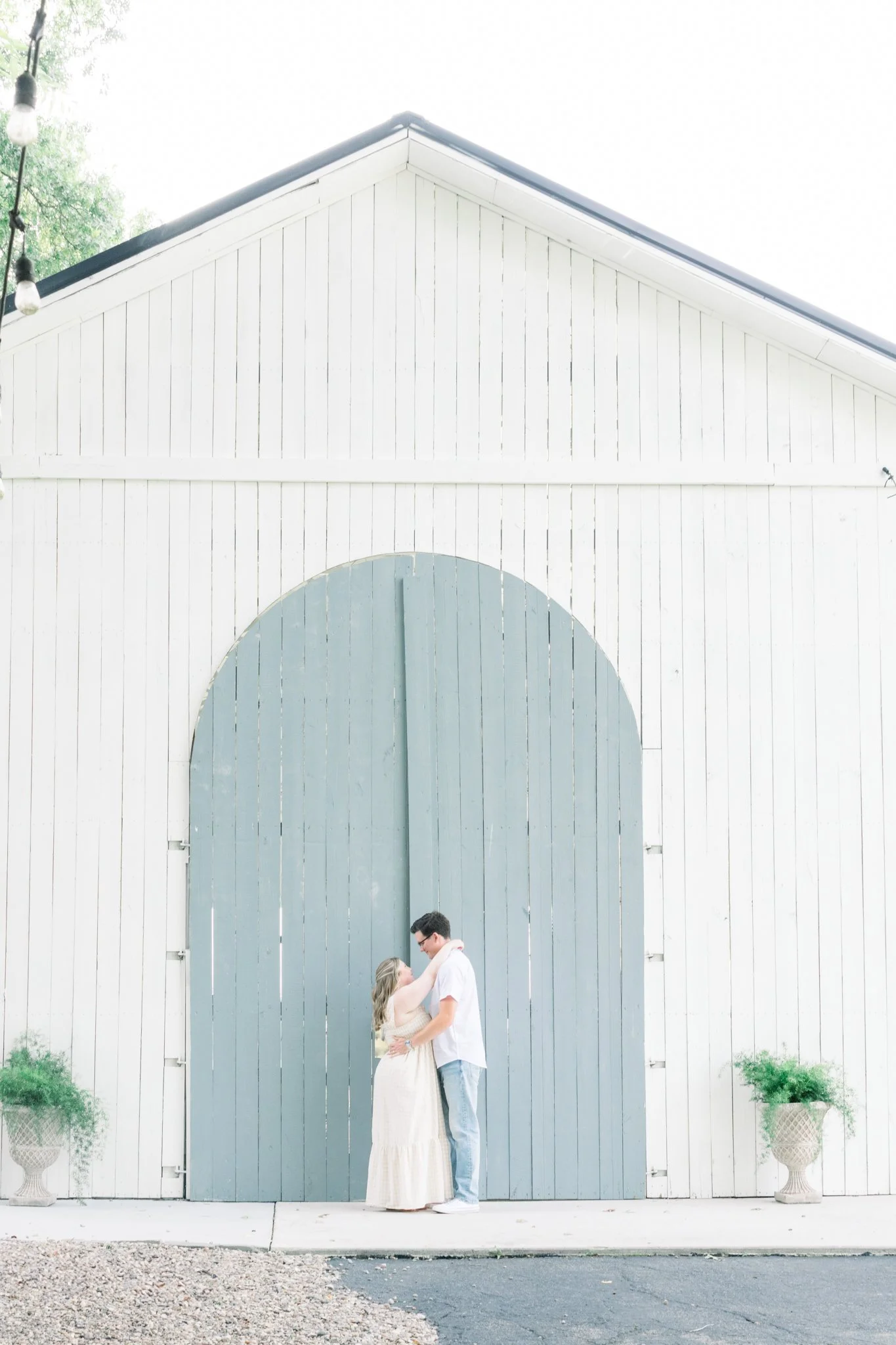 A couple stands close together in front of a large white barn door, embracing and gazing into each other's eyes. The woman wears a long beige dress and the man wears a white shirt and blue jeans. There are two potted plants on either side of the barn