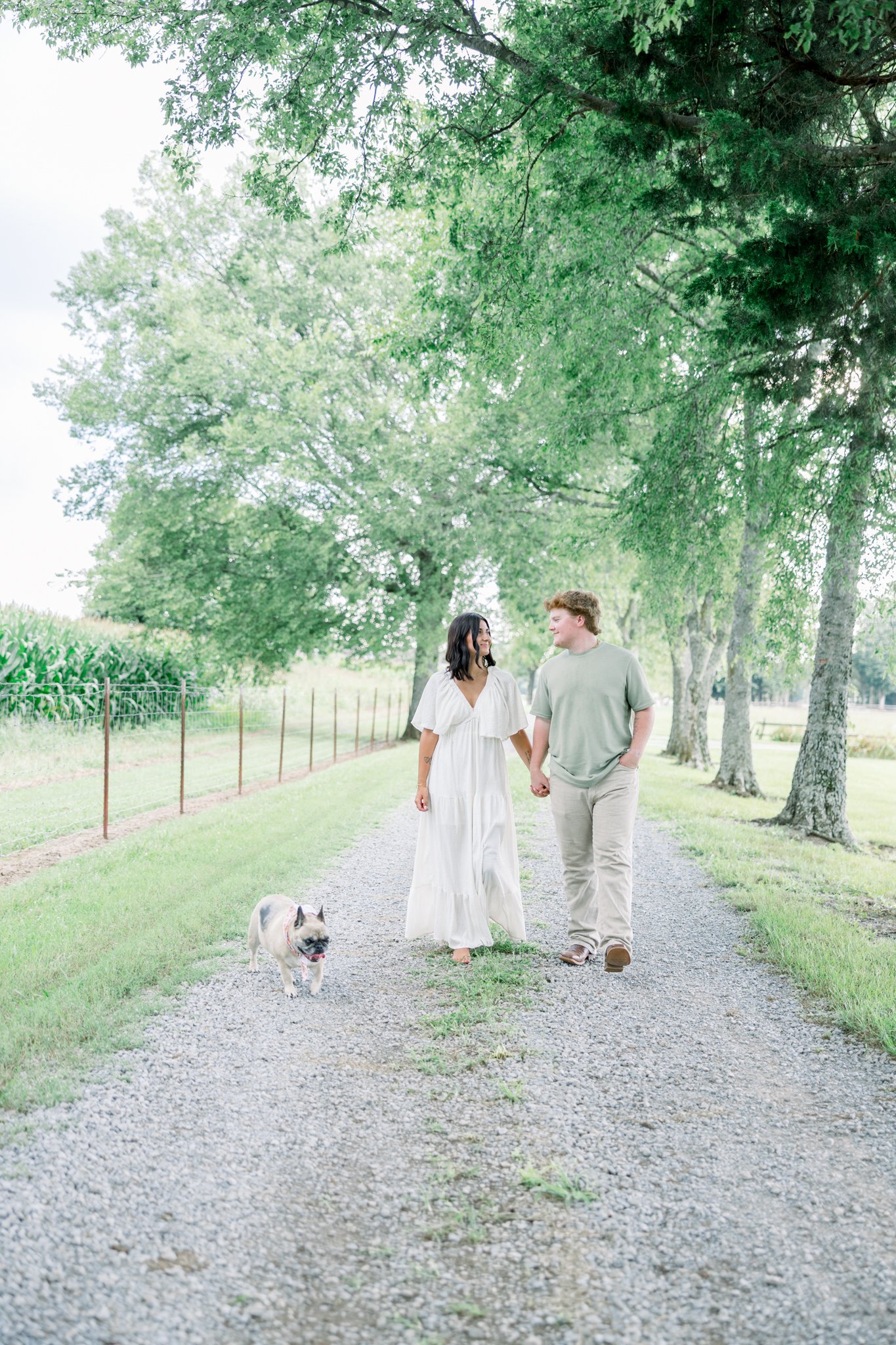 A couple walking hand in hand on a gravel path in a park with green trees and a dog. The woman is wearing a white dress, and the man is wearing a light green t-shirt and beige pants. The dog is small and light-colored with a dark face.