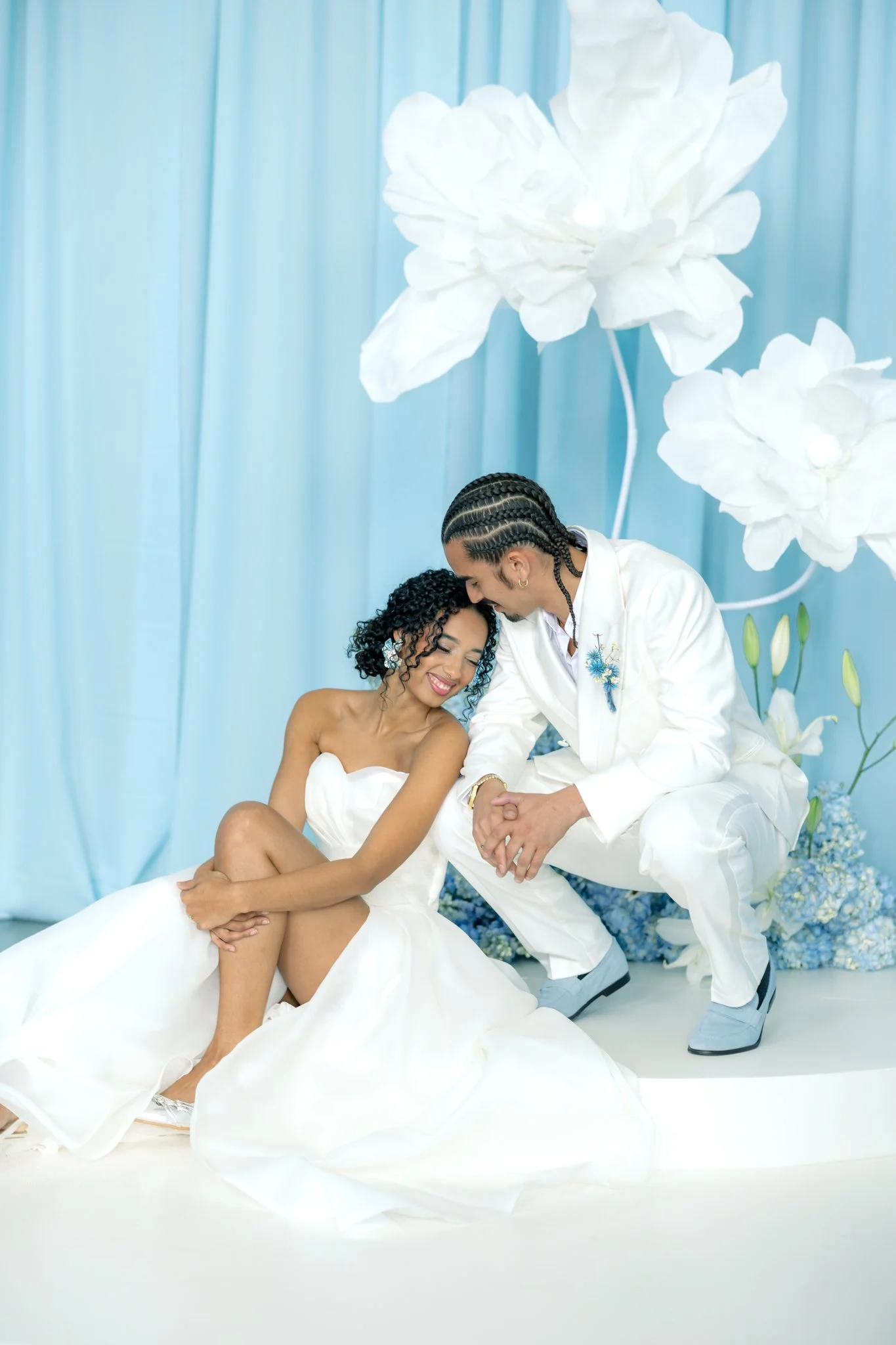 A couple in wedding attire with the bride sitting on the floor and the groom kneeling beside her, holding her hands, in front of a light blue backdrop with large white paper flowers.
