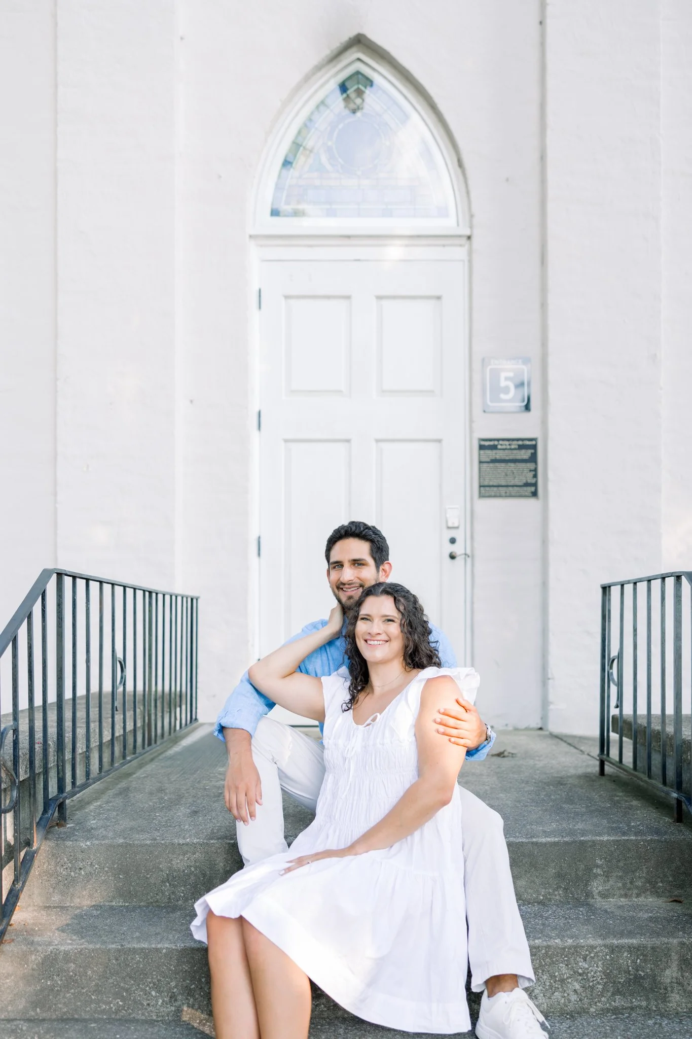 A smiling man and woman sitting on outdoor steps in front of a white church door, with a metal railing on either side, behind them.