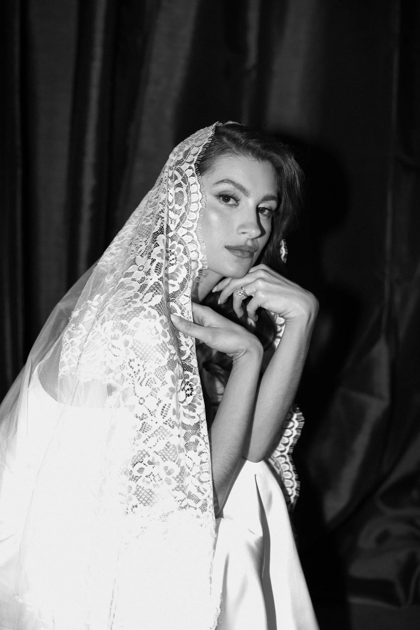 Black-and-white photo of a woman smiling, wearing a lace veil and elegant attire, with her hand resting on her chin, in front of a dark curtain