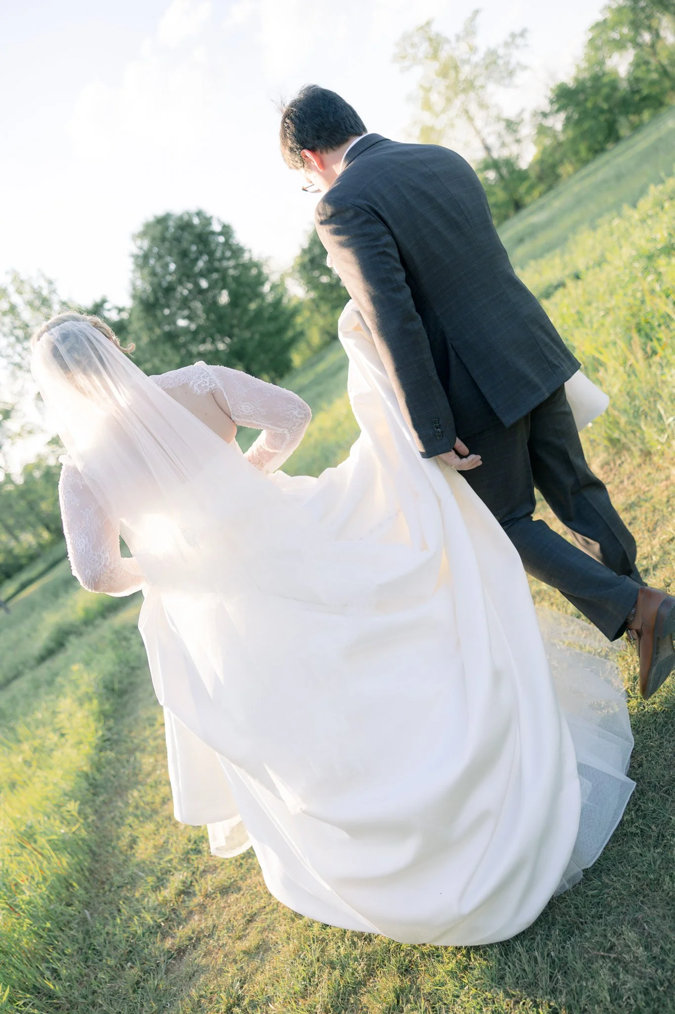 Wedding couple outdoors, woman in wedding dress with veil, man in dark suit, walking on grassy path with trees in background, late afternoon sunlight.