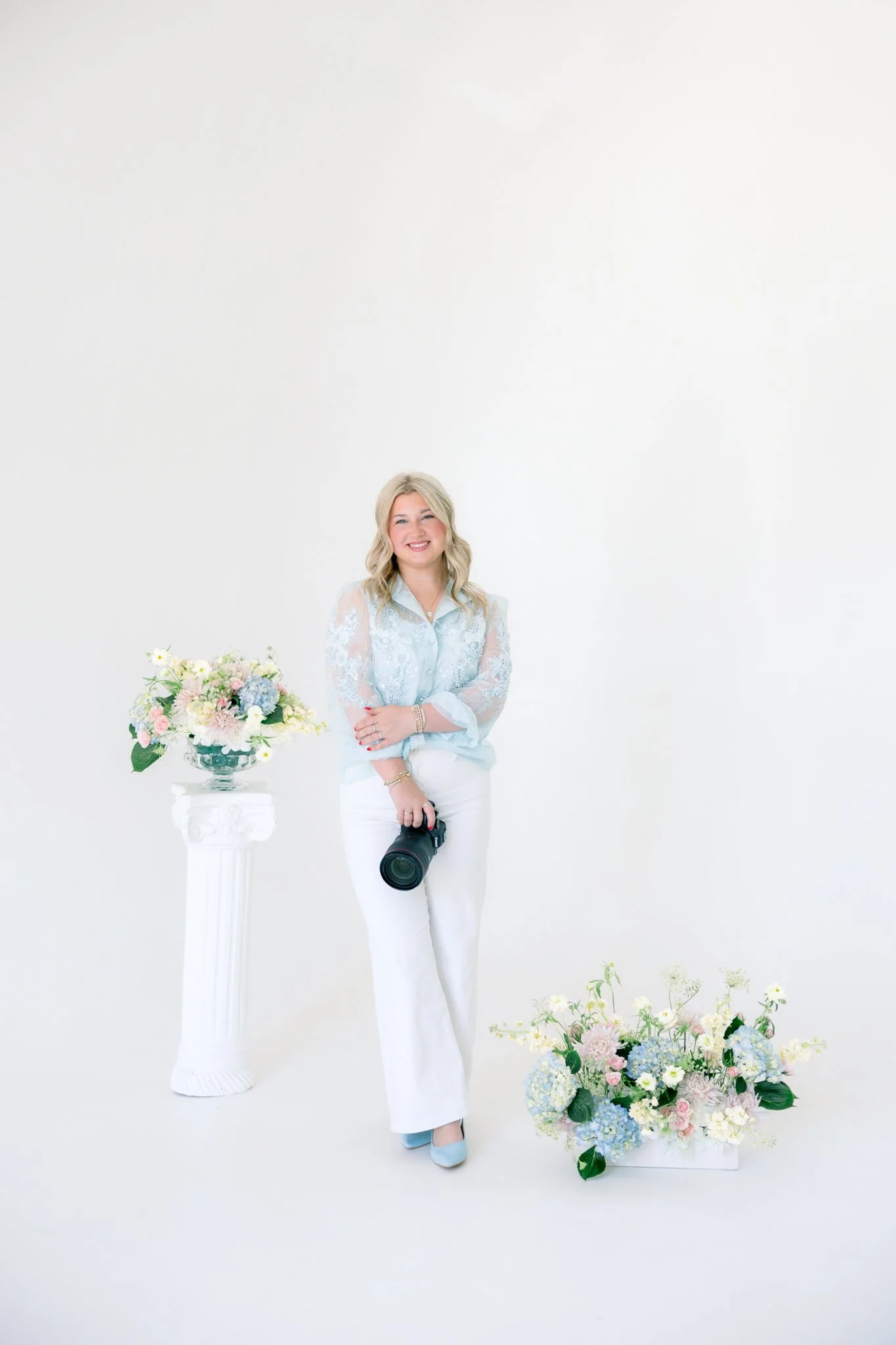 A woman with blonde hair smiling, wearing a light blue blouse with lace sleeves and white pants, stands beside a desk with a large floral arrangement in a white vase on a white pedestal, and another floral arrangement on the floor beside her, all against a plain white background.