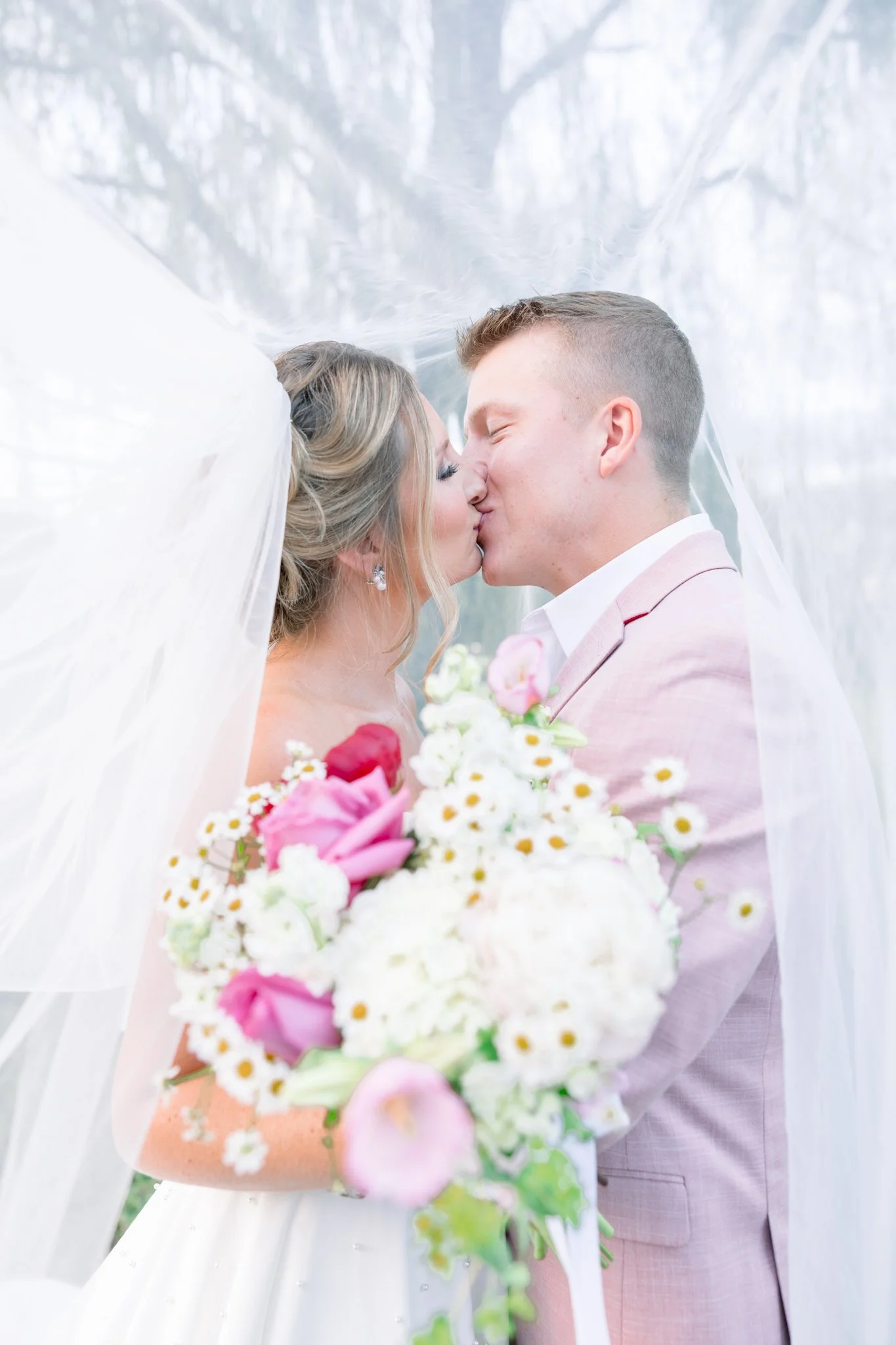 A bride and groom sharing a kiss during their wedding ceremony, with the bride holding a bouquet of pink and white flowers, surrounded by a white veil and softly blurred outdoor background.