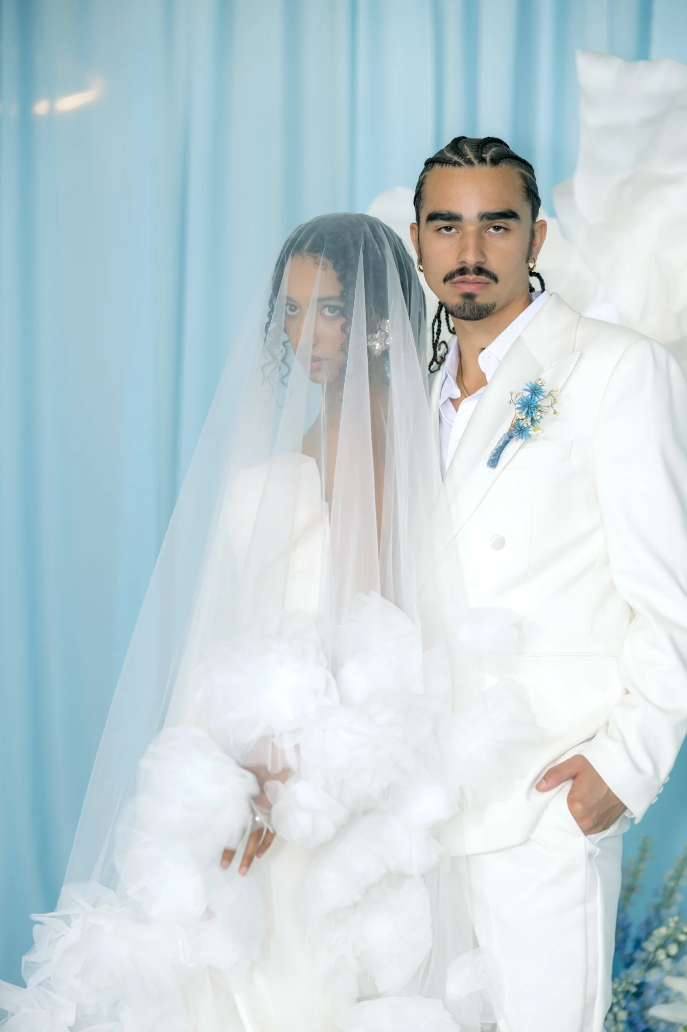 A man and a woman dressed in wedding attire, with the woman wearing a veil and the man in a white suit, standing together in front of a light blue background with soft lighting.