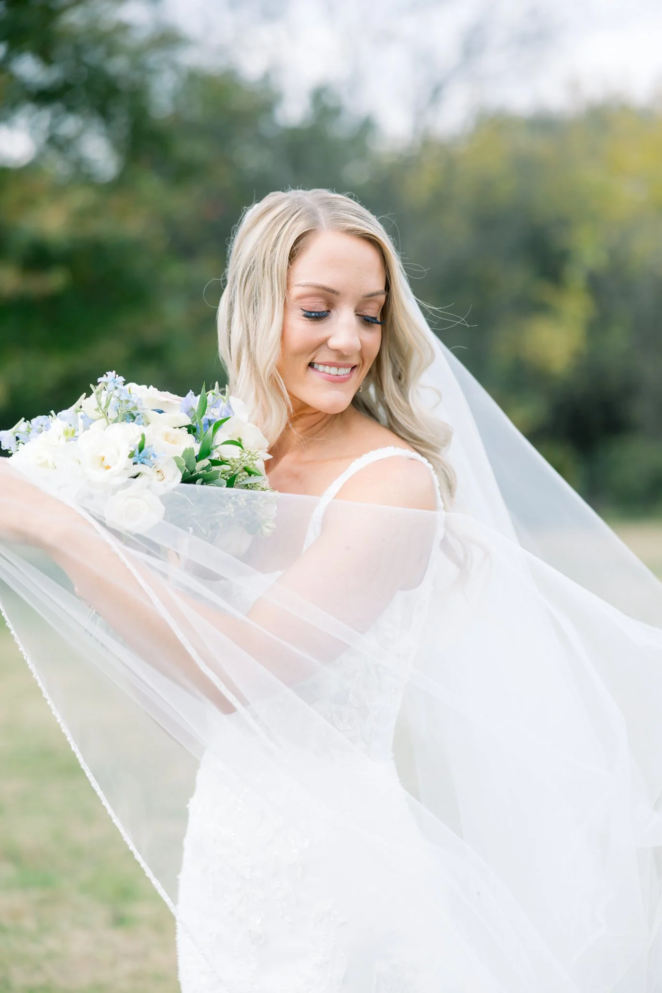 A smiling bride with blonde hair and blue eye makeup, holding a bouquet of white and purple flowers outdoors.