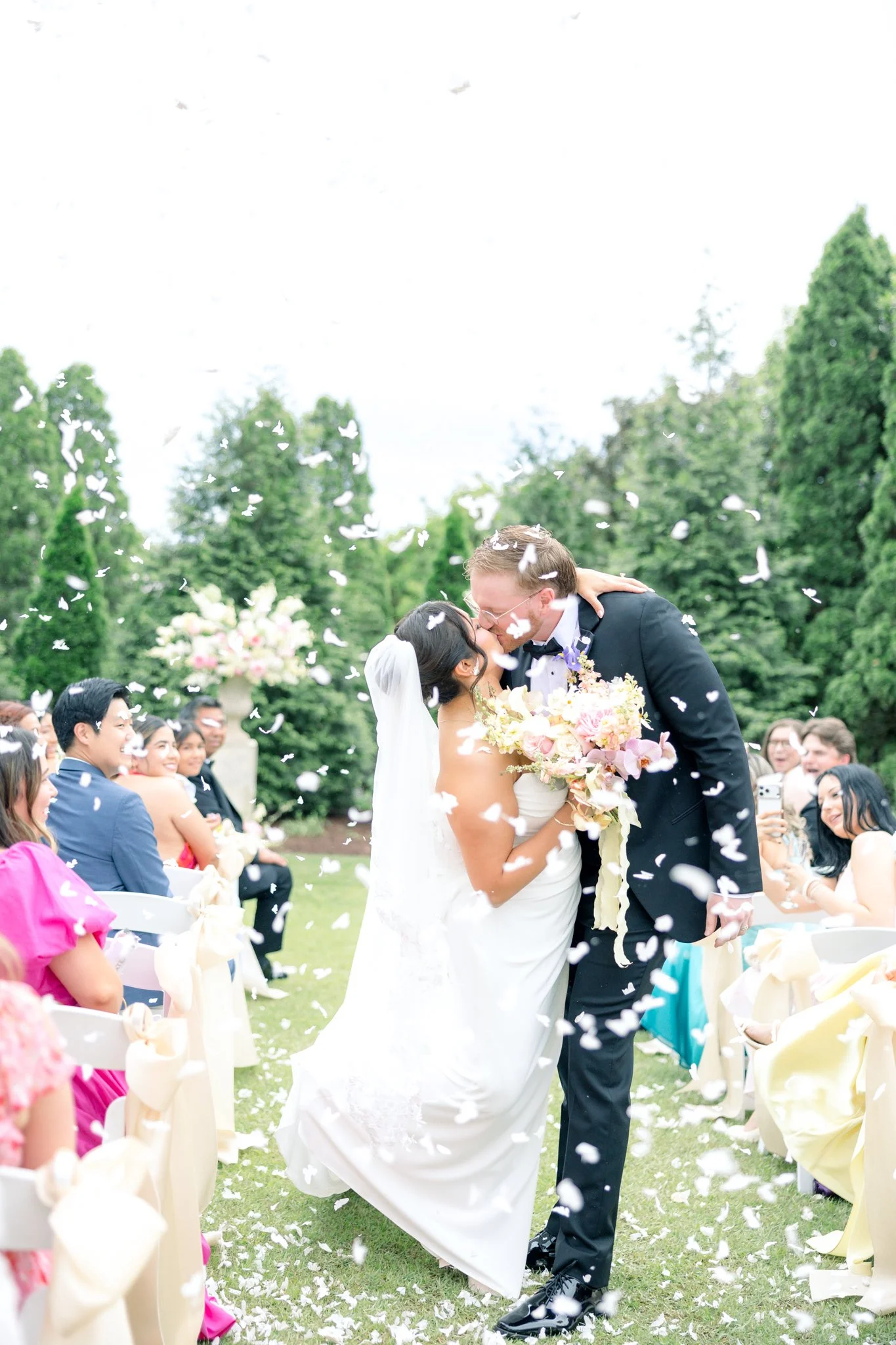 A bride and groom kiss during their outdoor wedding ceremony, surrounded by seated guests, with flower petals falling around them and green trees in the background.