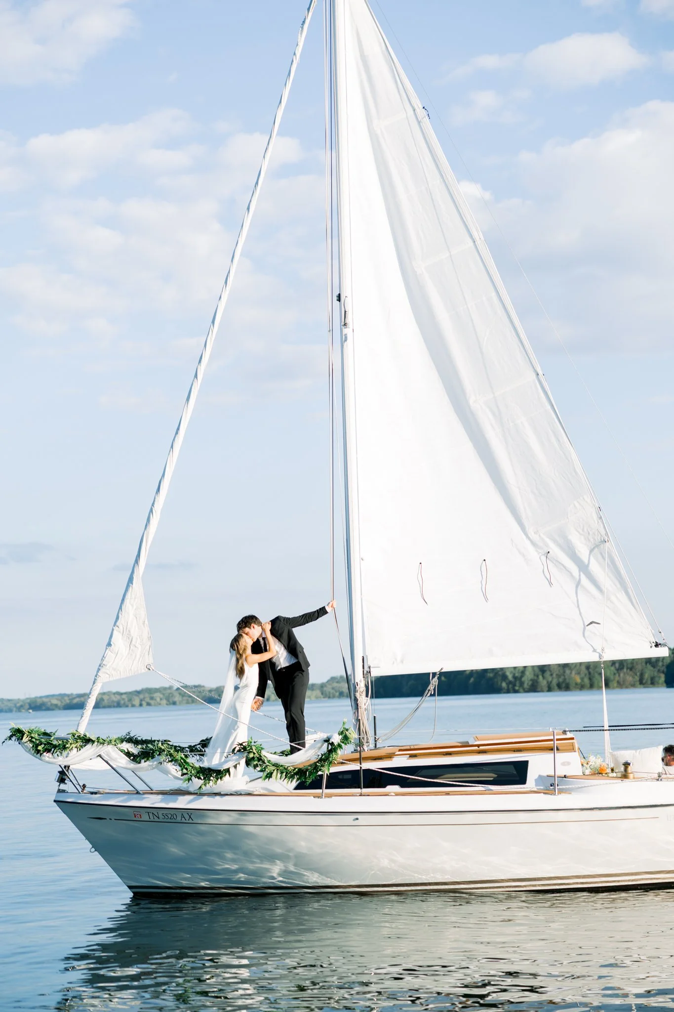 A couple dressed in wedding attire, standing on a sailboat decorated with greenery, sharing a kiss while the boat is in the water during daytime.