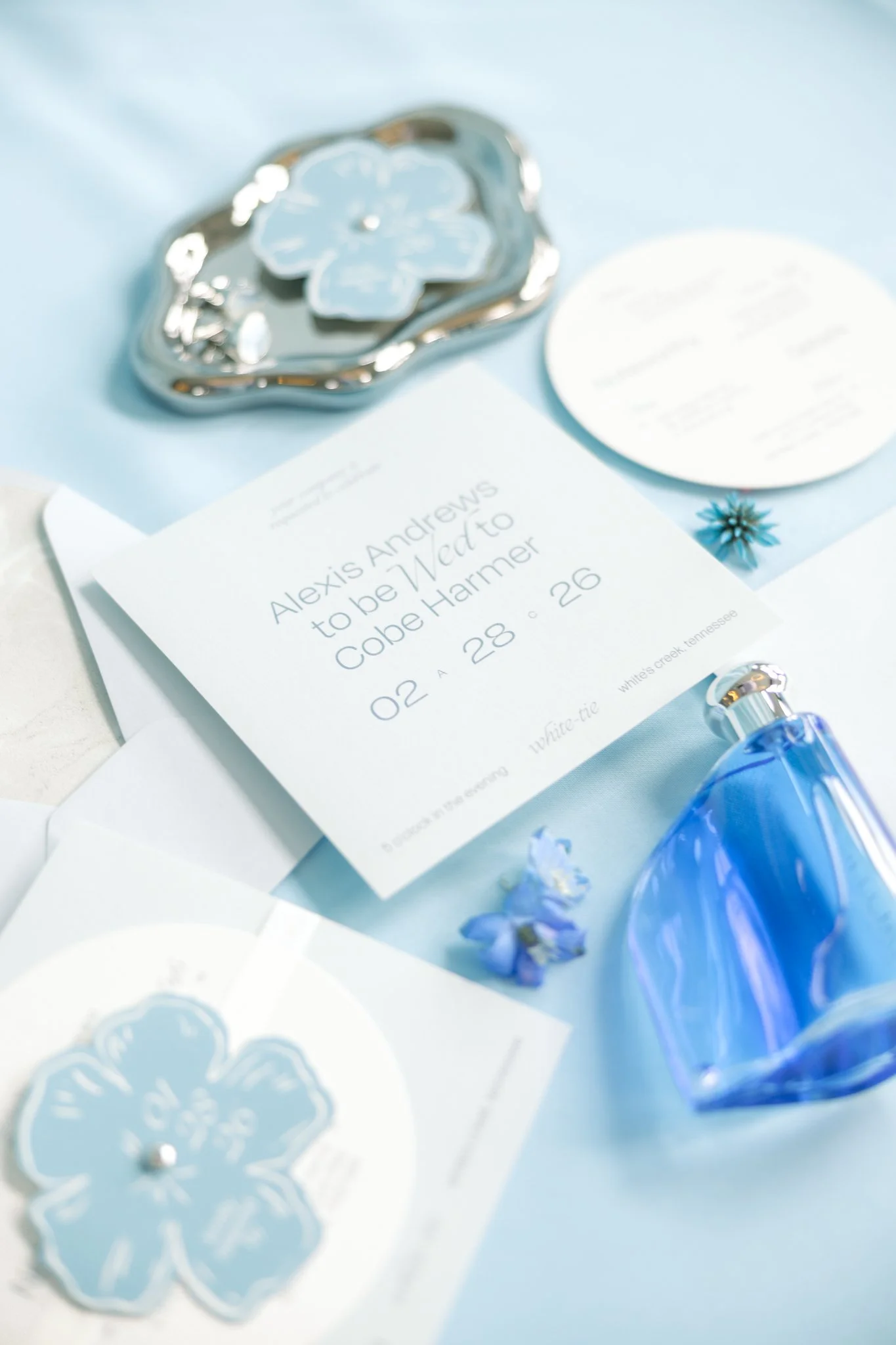 Invitation card for Alexis Andrews' wedding, scheduled for February 28, 2026, in White's Creek, Tennessee, along with some blue floral decorations, a blue perfume bottle, and decorative stickers featuring blue flowers.