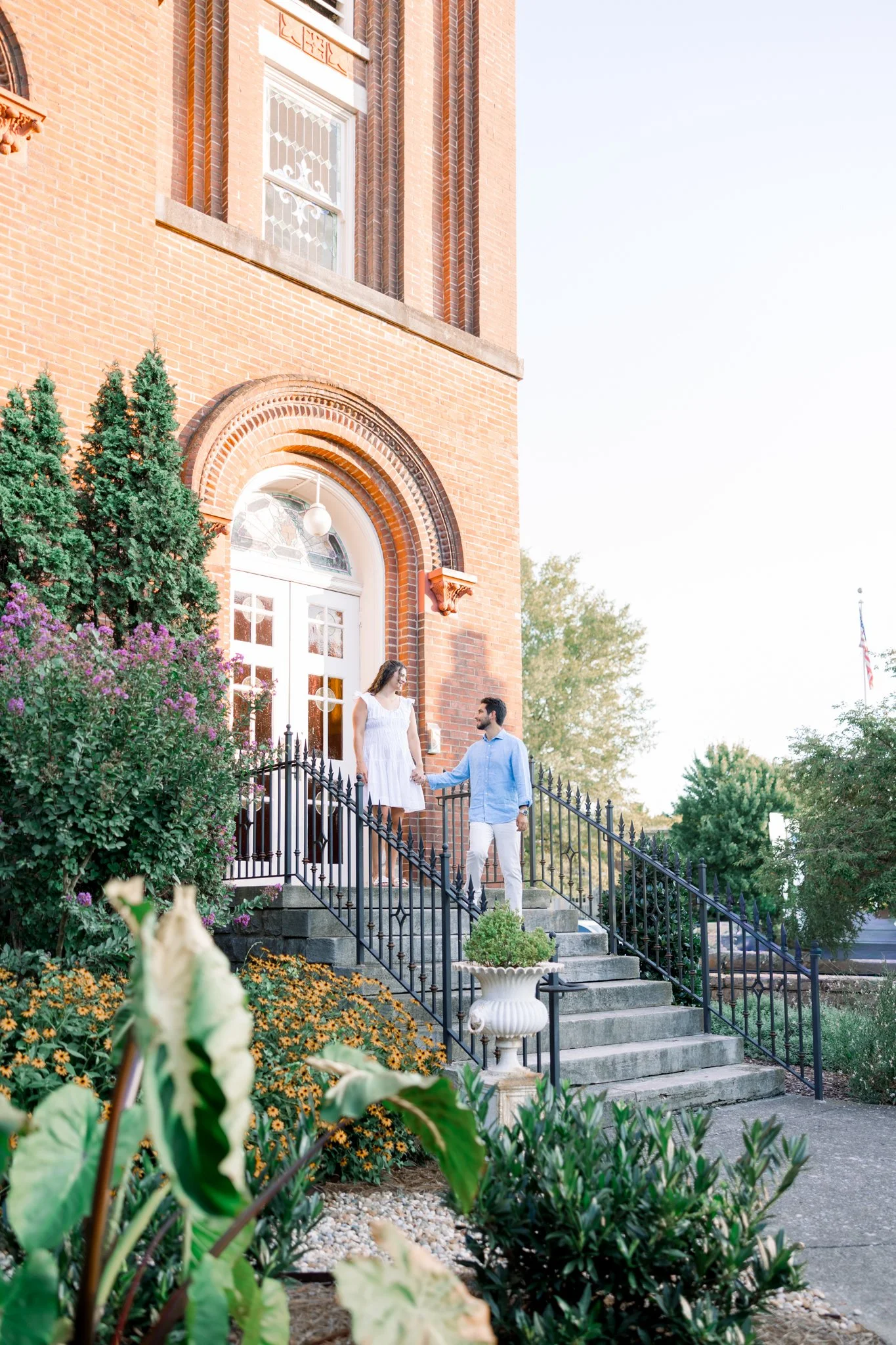 A couple is standing on the steps of a brick building, holding hands. The woman is wearing a white dress, and the man is wearing a light blue shirt and white pants. There are green shrubs and colorful flowers surrounding the stairs, and the building has decorative architectural details around the door and windows.