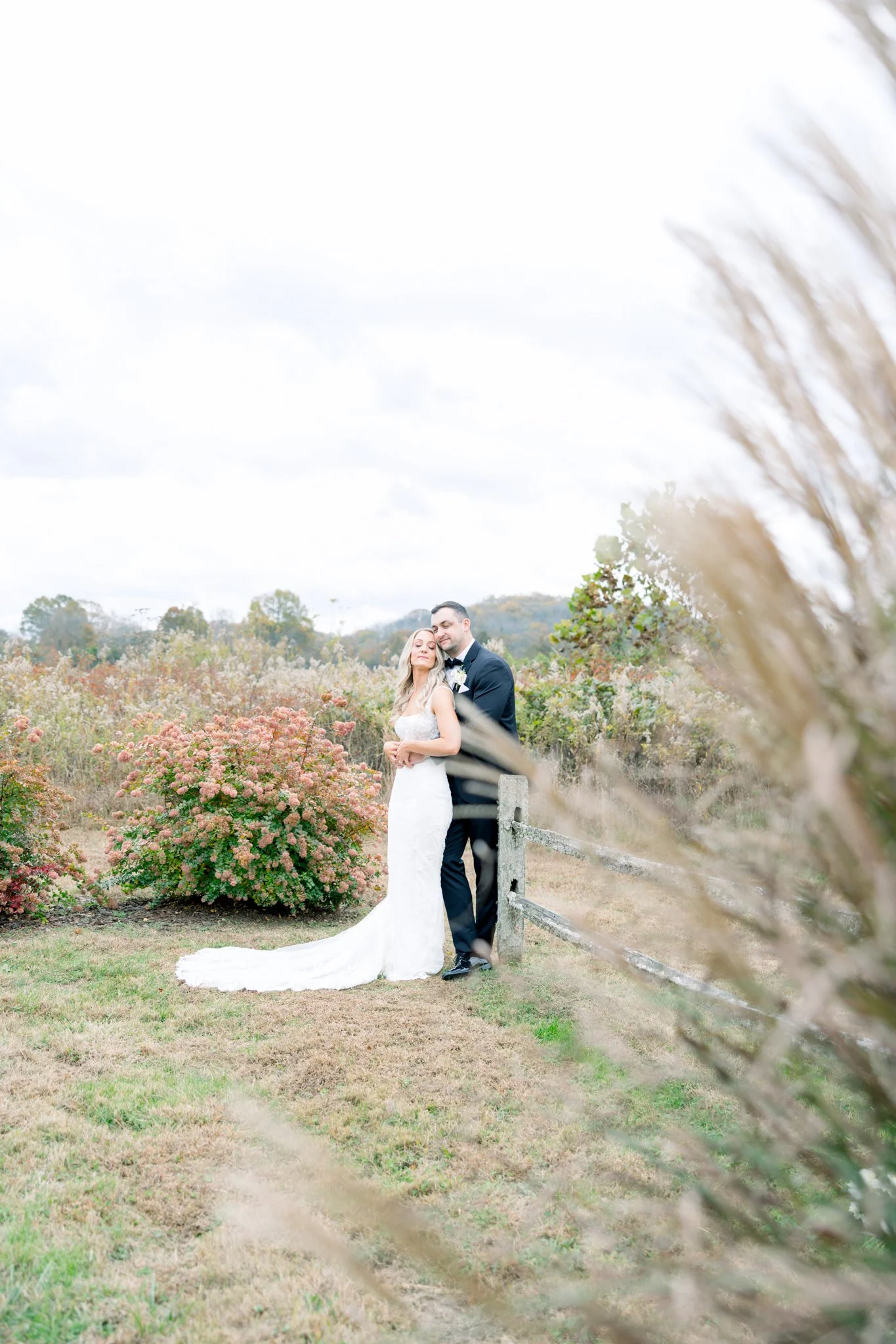 A bride and groom standing close together outdoors in a scenic field with flowers and trees, with a rustic fence visible. The bride is wearing a white wedding gown, and the groom is in a dark suit. The couple looks content, sharing an intimate moment
