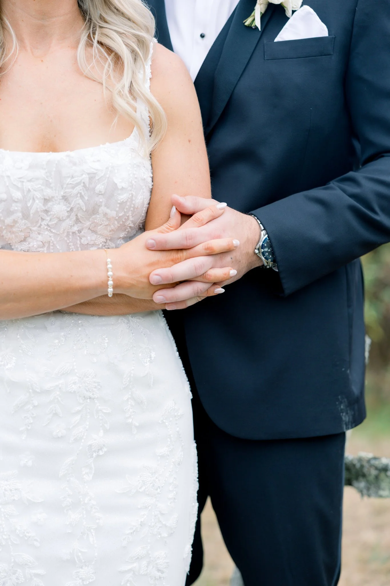 Close-up of a bride and groom holding hands during a wedding ceremony, with the bride wearing a white lace wedding dress and a pearl bracelet, and the groom wearing a dark suit with a white shirt, a boutonniere, and a watch.