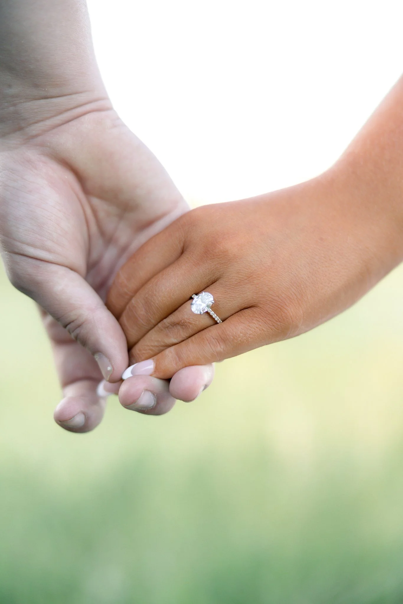 Close-up of a woman wearing a diamond engagement ring holding hands with another person, against a soft, blurred yellow-green background.