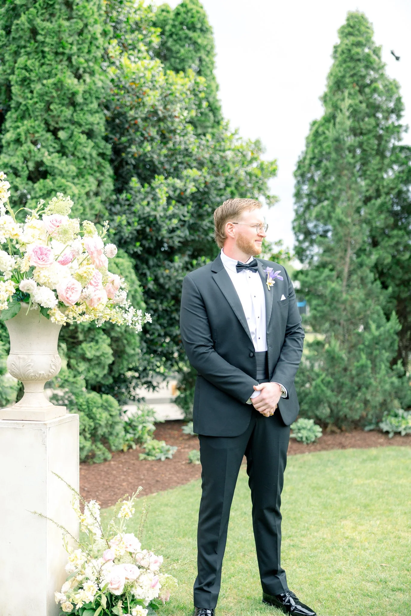 A man in a black tuxedo and bow tie standing outdoors near a large flower arrangement.