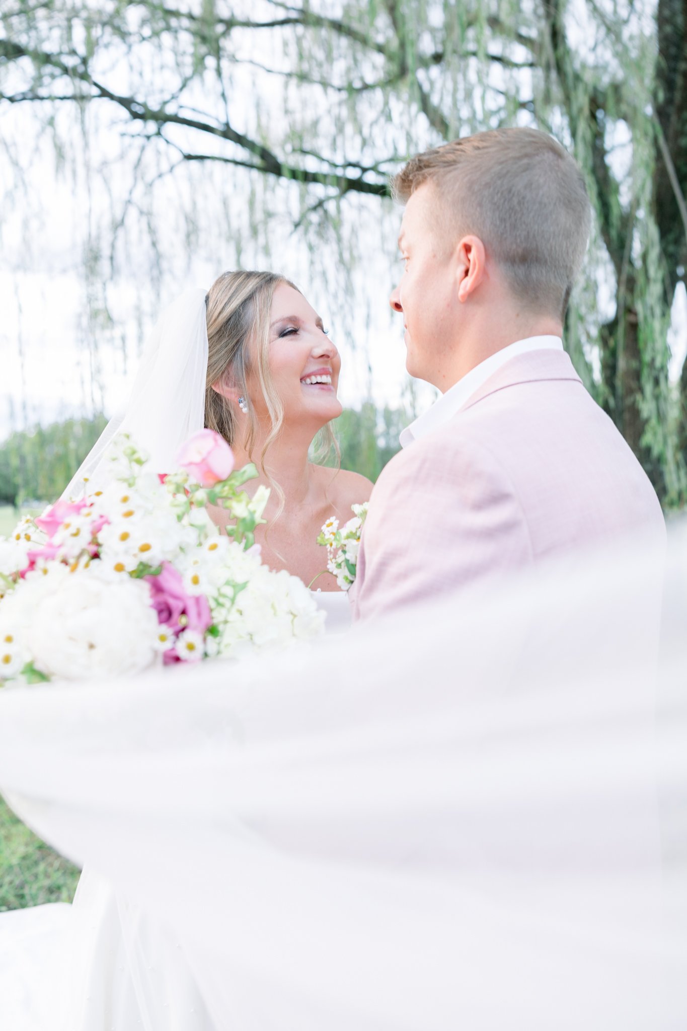 A bride and groom smiling and looking at each other during their outdoor wedding ceremony, with a bouquet of pink and white flowers in the foreground and trees in the background.
